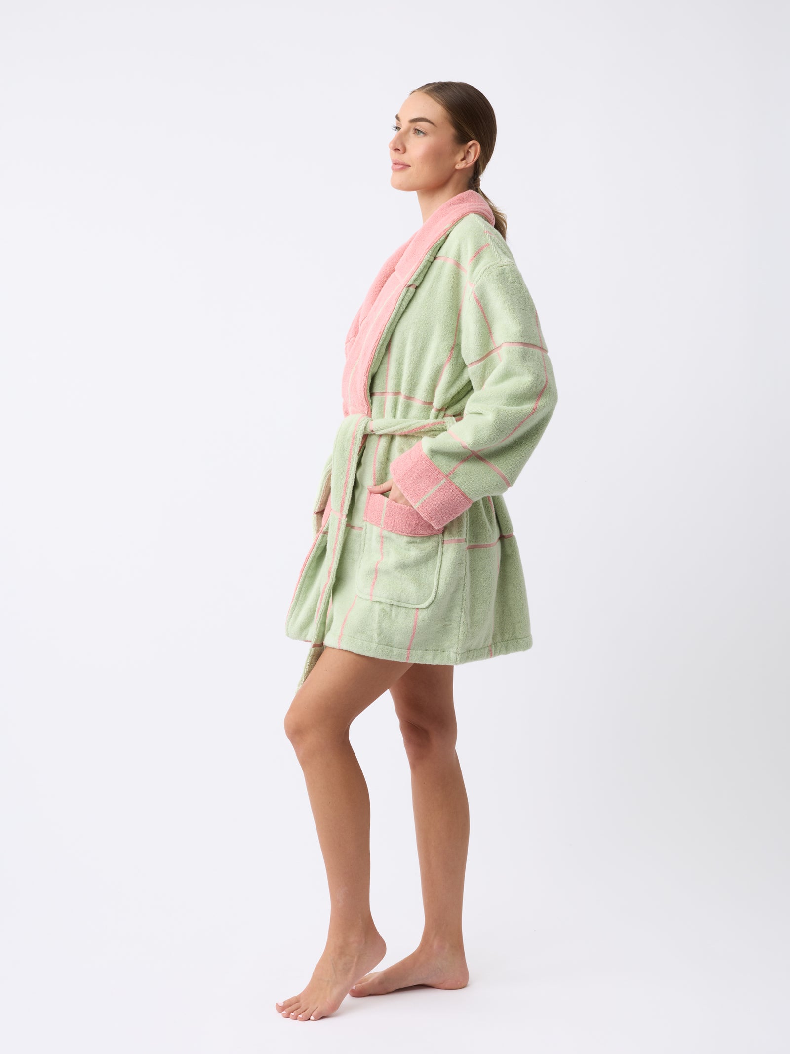 A person stands barefoot on a white background wearing the Cozy Earth Windowpane Resort Robe, featuring pastel green and pink checks with a belt. The robe has pink cuffs and a collar, and the person is slightly turned to the side, looking forward. 