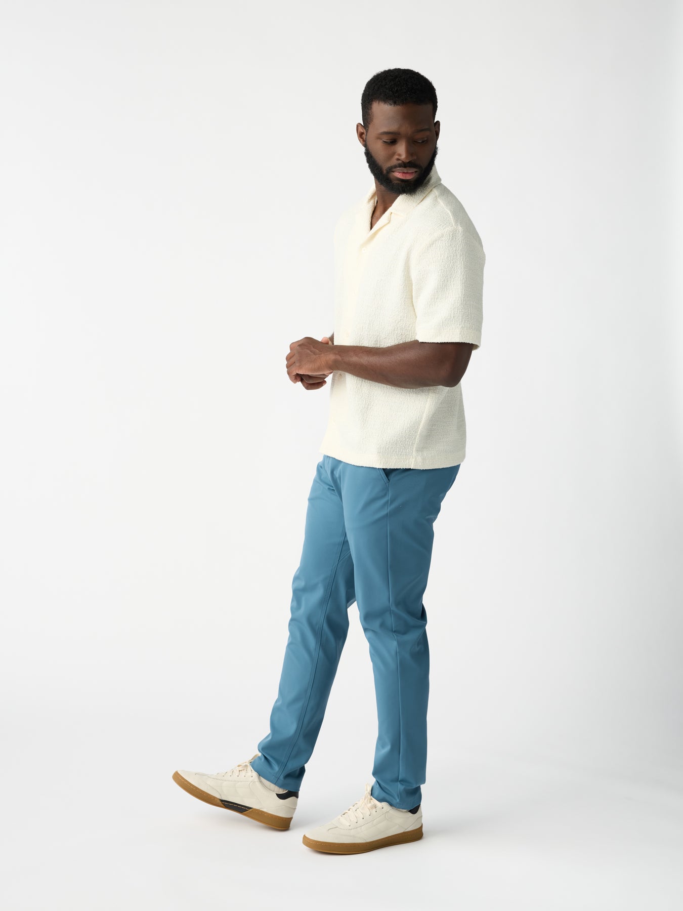 A man poses against a plain white background, wearing a textured short-sleeve white shirt, Cozy Earth's Men's Everywhere Pant in light blue, and cream sneakers with tan soles. He sports a beard and looks to the side. 