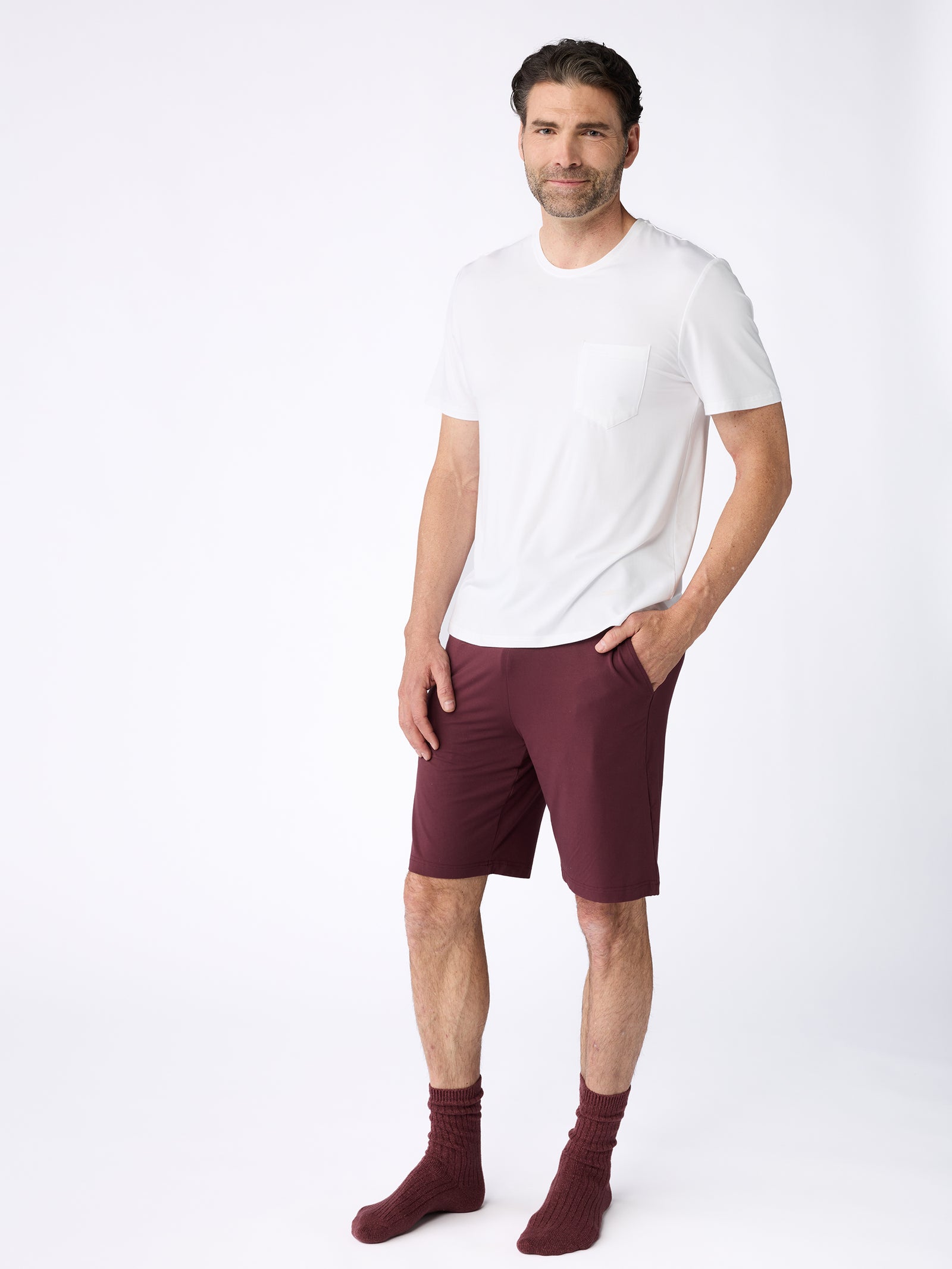 A man stands against a plain white background, wearing a Lounge Tee from Cozy Earth, maroon shorts, and maroon socks. His hands are in his pockets, and he is smiling slightly. 