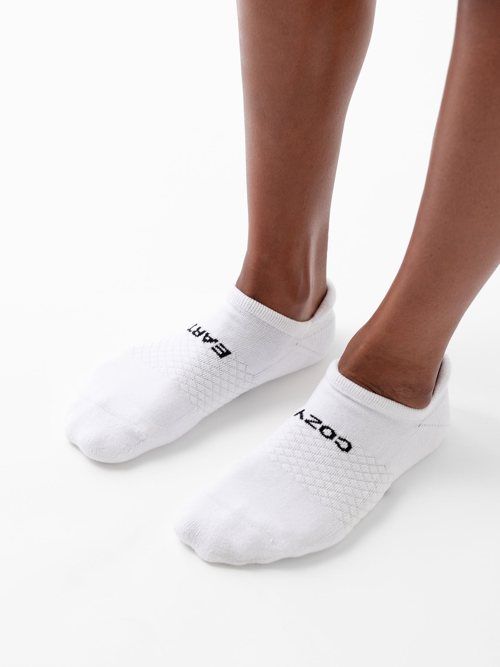 Close-up of a person's lower legs and feet wearing white athletic ankle socks from the Essential Ankle Sock 2-Pack by Cozy Earth. The left sock features the text "EART" near the cuff, while the right sock displays "COZY." The background is plain white. 