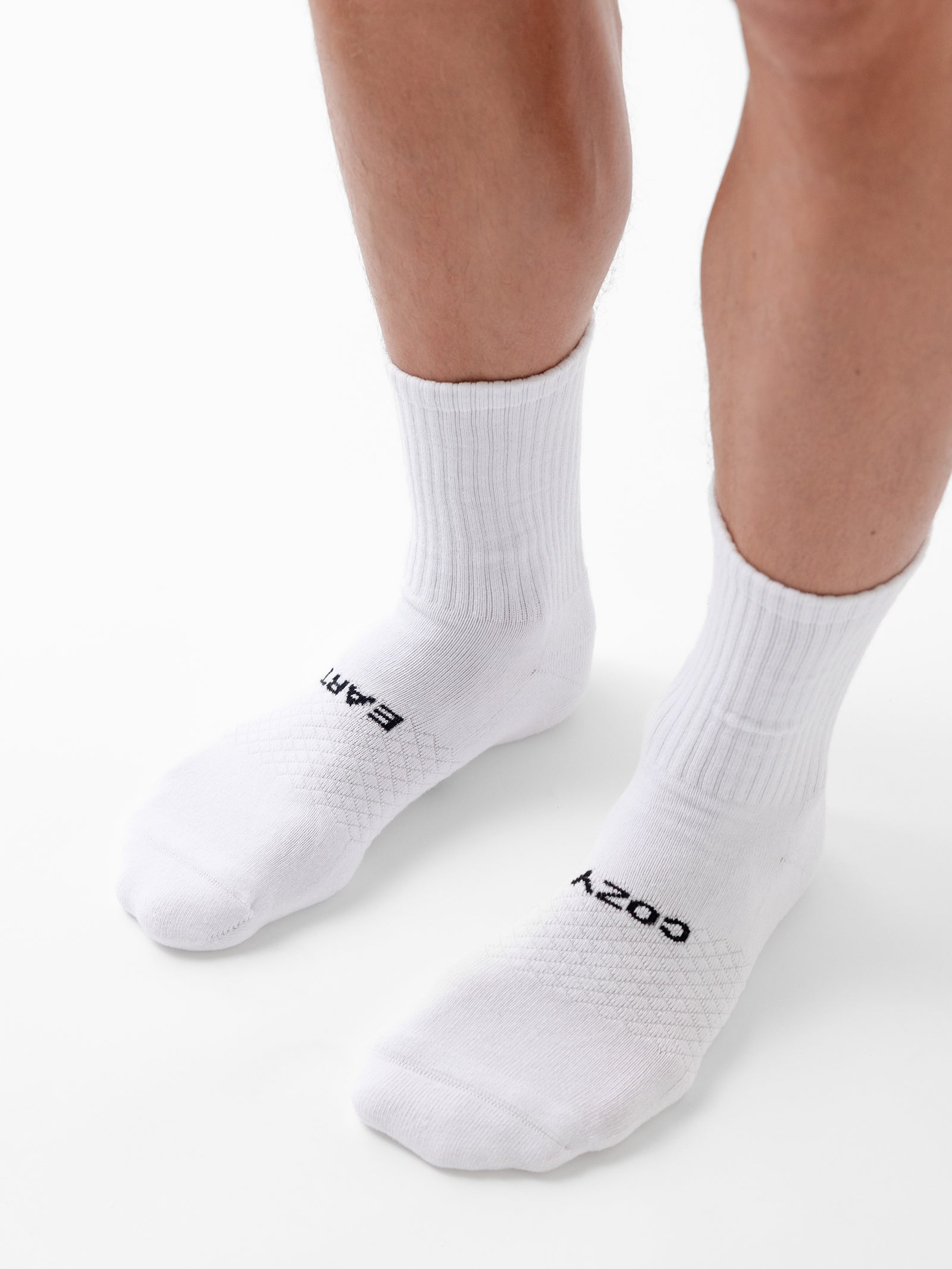 Close-up of a person's lower legs and feet wearing white athletic socks from Cozy Earth's Essential Calf Sock 4-Pack. The socks feature the words "EAR" and "COZY" written on them in black text. The person stands on a white surface. 