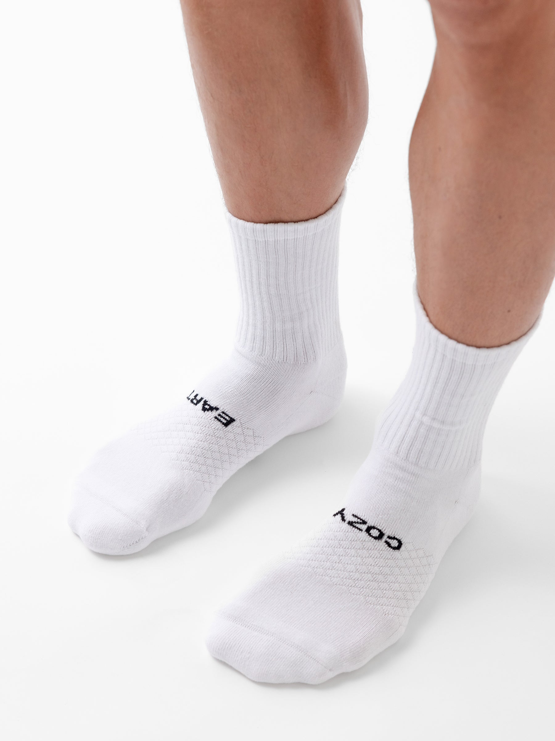 A person wears white Essential Calf Socks from Cozy Earth; the left sock reads "EARTH" and the right "COZY" in black letters. The background is plain white. 
