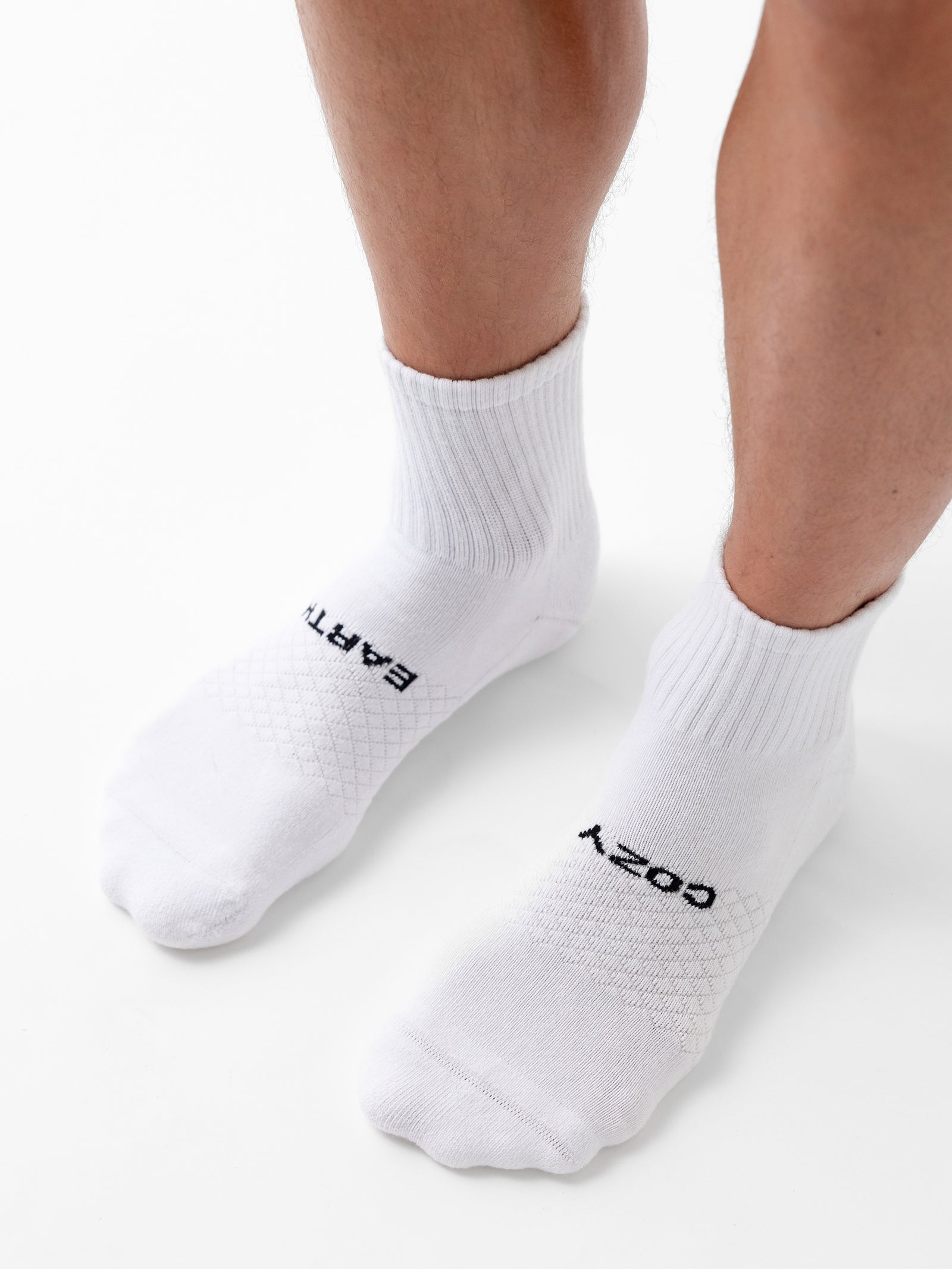 A pair of feet wearing white athletic socks from the Cozy Earth Essential Quarter Sock 2-Pack, standing on a white surface. The socks have the words "EARTH" and "COZY" printed on top near the toes, and they feature ribbed cuffs along with a textured pattern on the top. 