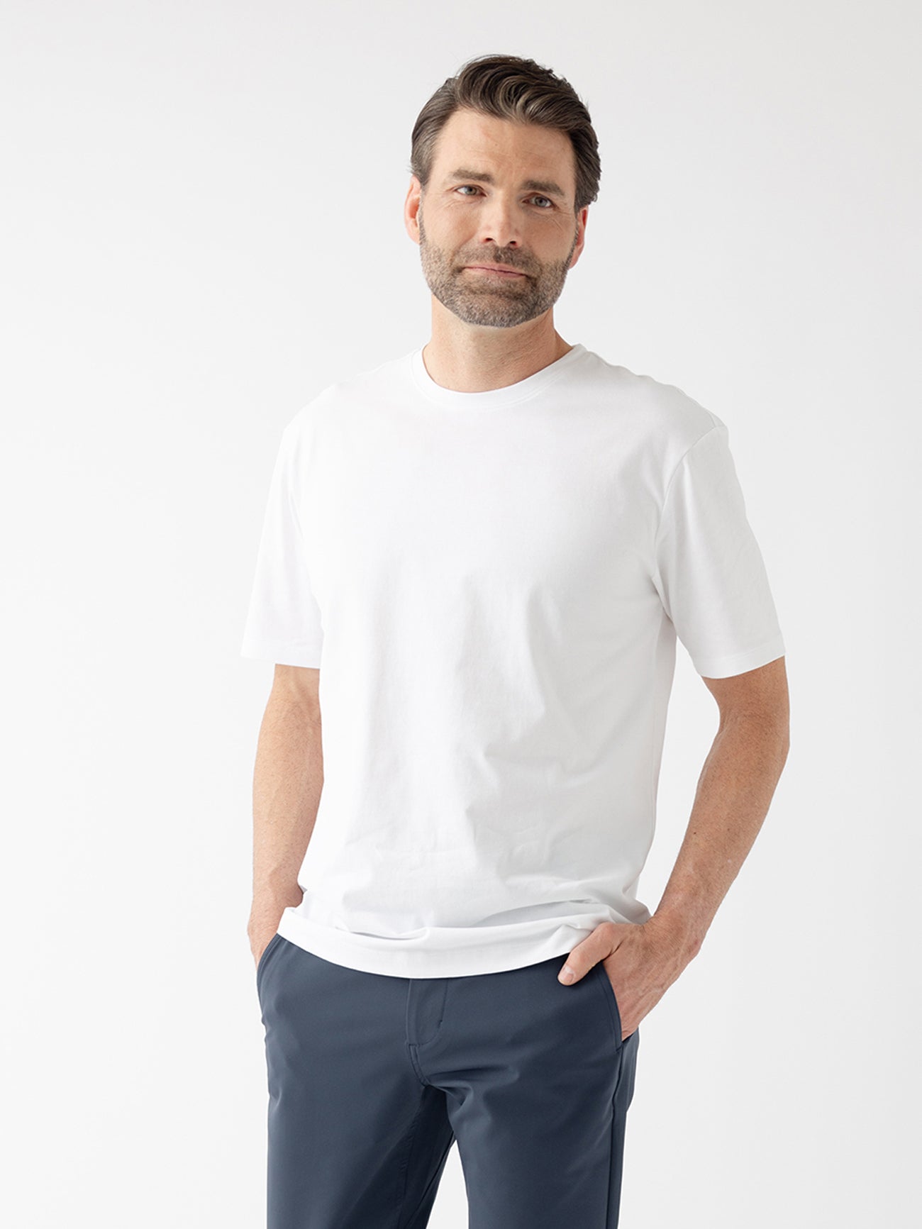 A man with short dark hair and a beard stands against a plain white background, wearing the Cozy Earth Men's All Day Tee and dark pants. His hands are in his pockets as he looks towards the camera with a neutral expression. |Color:White