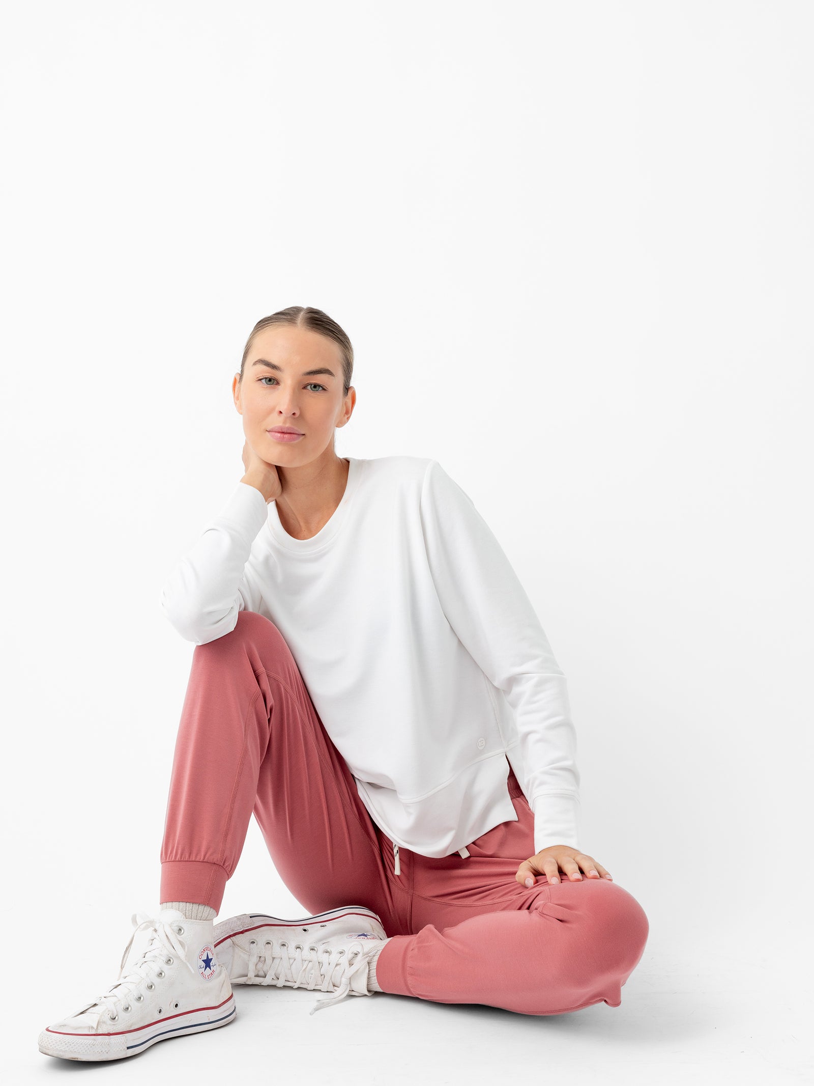 A person with long, dark hair tied back sits casually on the floor against a white background. They are wearing a Cozy Earth Women's StudioLite Crewneck, pink sweatpants, and white high-top sneakers featuring a blue and red star logo. One arm rests on their knee, and they have a relaxed expression. 