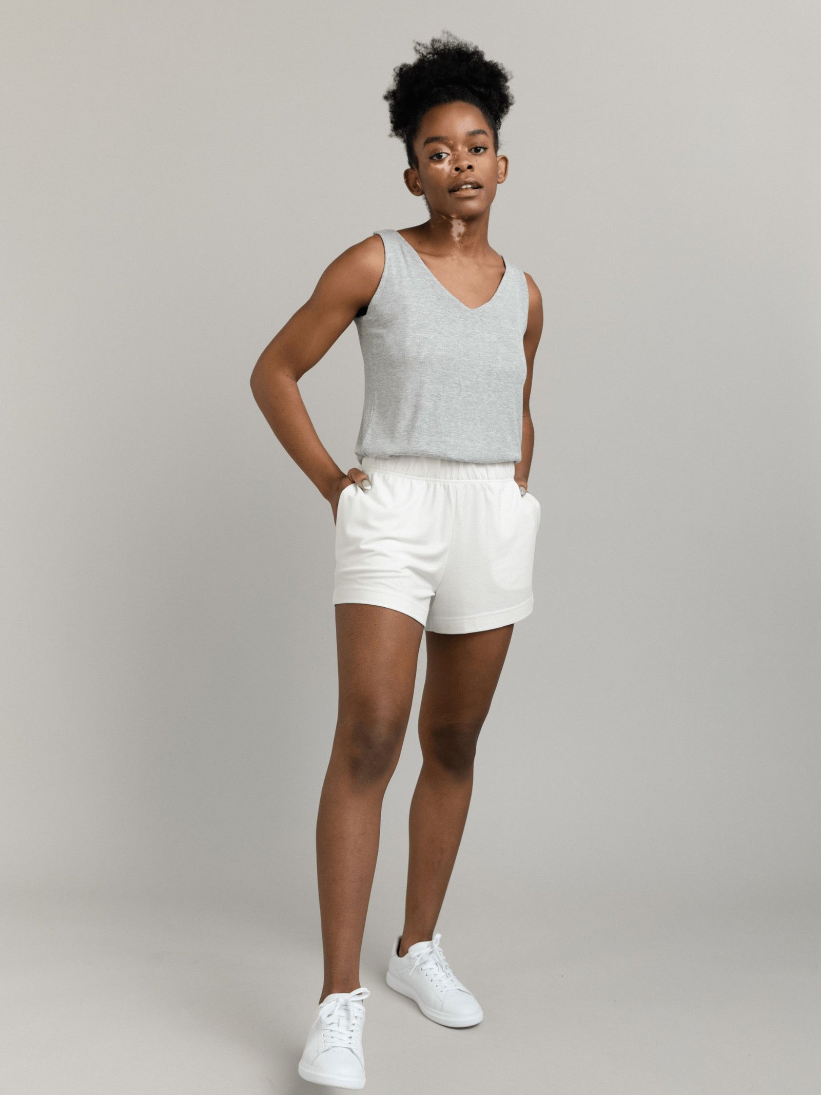 In a minimalist studio with a light gray background, a person confidently sports Cozy Earth's Women's Modern Modal Shorts in white, paired with a light gray sleeveless top and white sneakers, their short curly hair complementing their casual pose with hands in pockets.