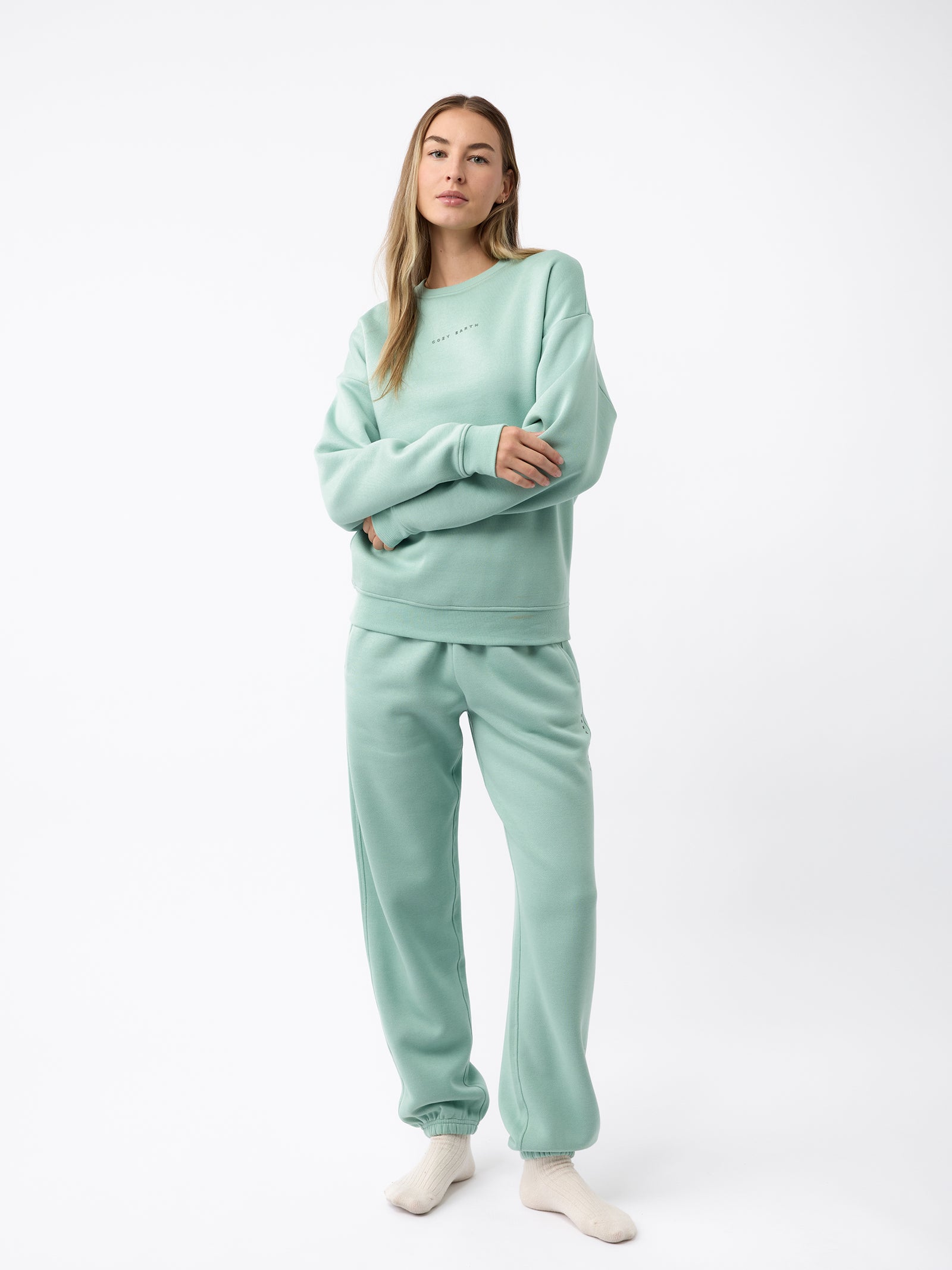 A woman wearing the Women's CityScape Crewneck by Cozy Earth stands gracefully in a light green sweatshirt and sweatpants. With long flowing hair and a direct gaze fixed on the camera, she embodies a casual and comfortable style, standing barefoot against a plain white backdrop. 