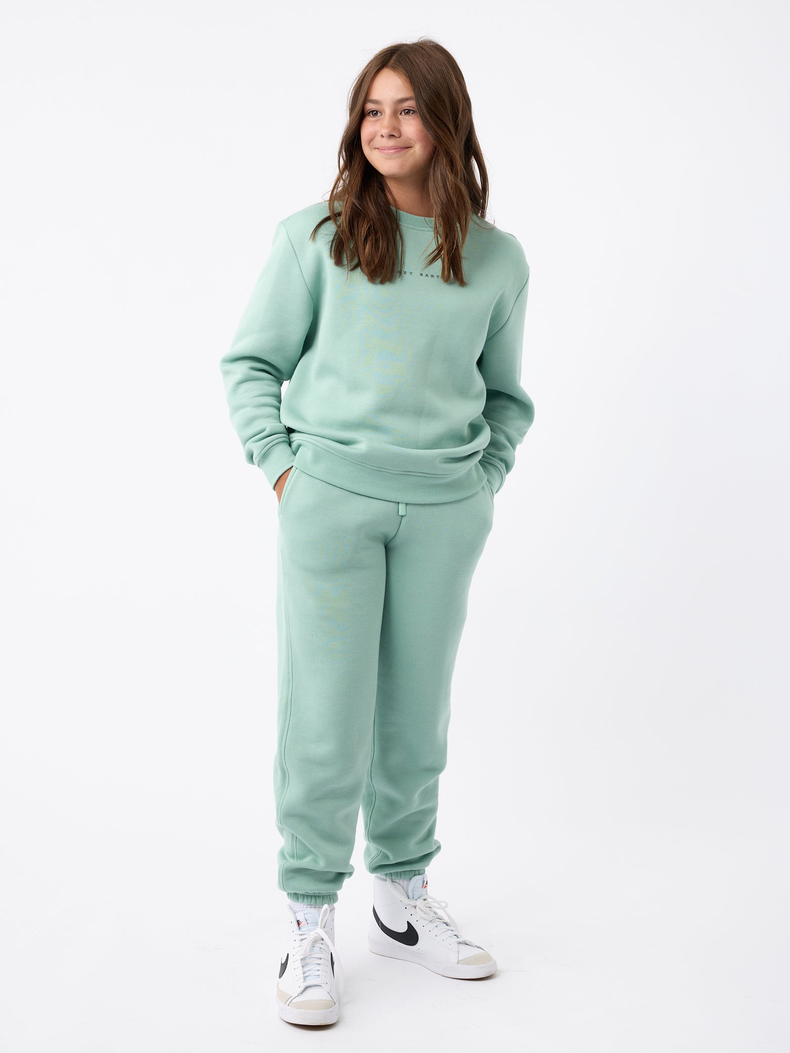 A person wearing Cozy Earth's Kid's CityScape Crewneck in mint green along with matching sweatpants stands against a plain white background. They have their hands in their pockets and are sporting white sneakers featuring black accents. 