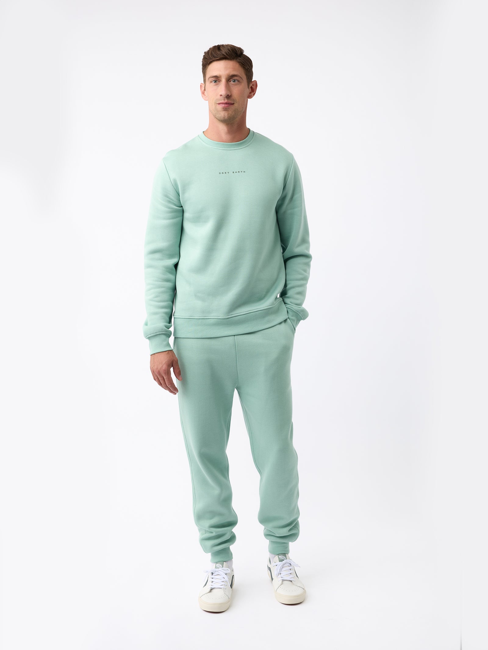 A person wearing the Cozy Earth's Men's CityScape Crewneck in light green, along with matching sweatpants, stands against a plain white background. They are also sporting white sneakers. The outfit looks casual and comfortable. 