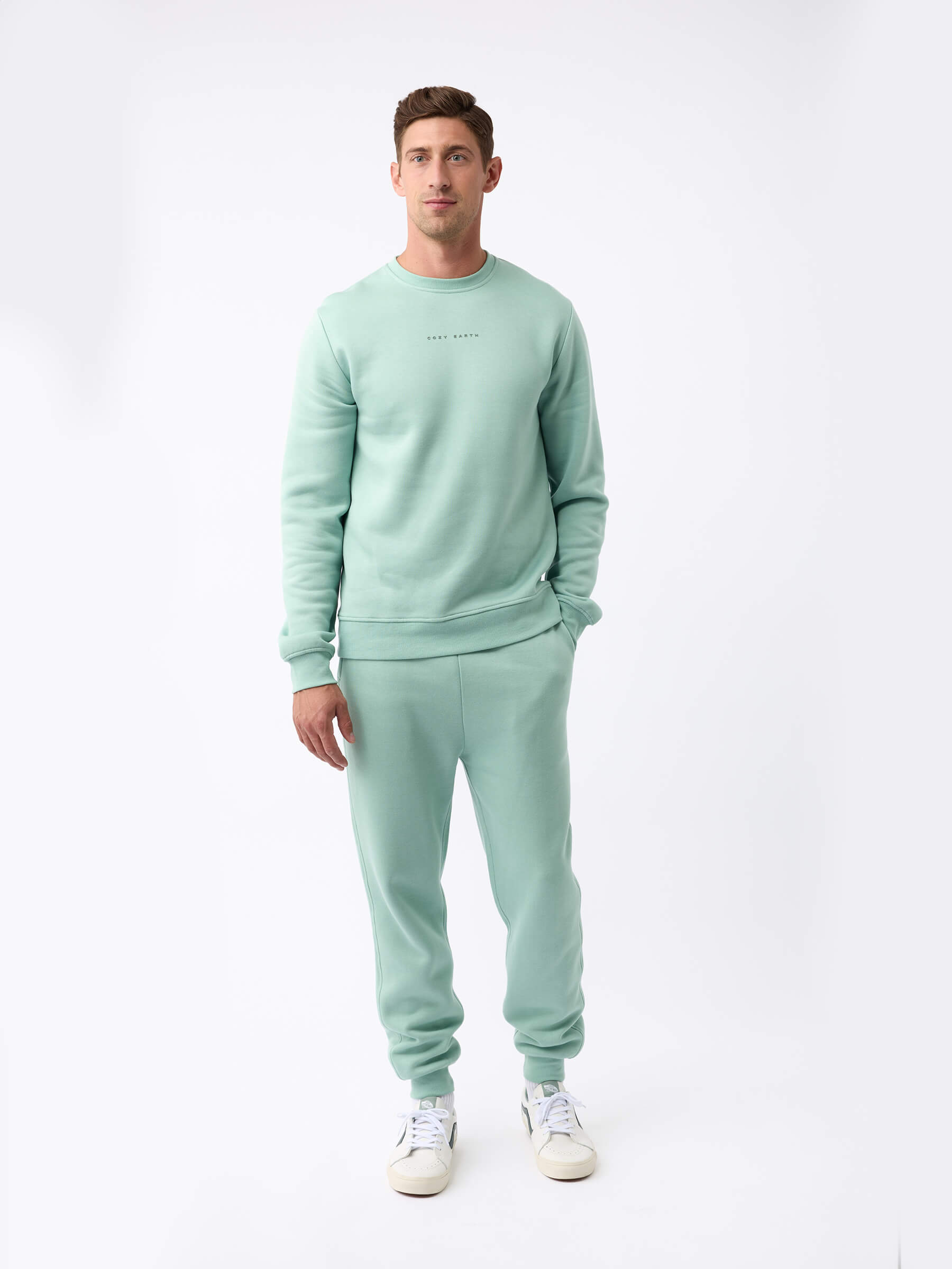 A man stands against a plain white background, wearing the Cozy Earth Men's CityScape Crewneck - Last Chance with matching sweatpants and white sneakers. His hands are relaxed by his sides and in his pocket as he looks slightly past the camera.