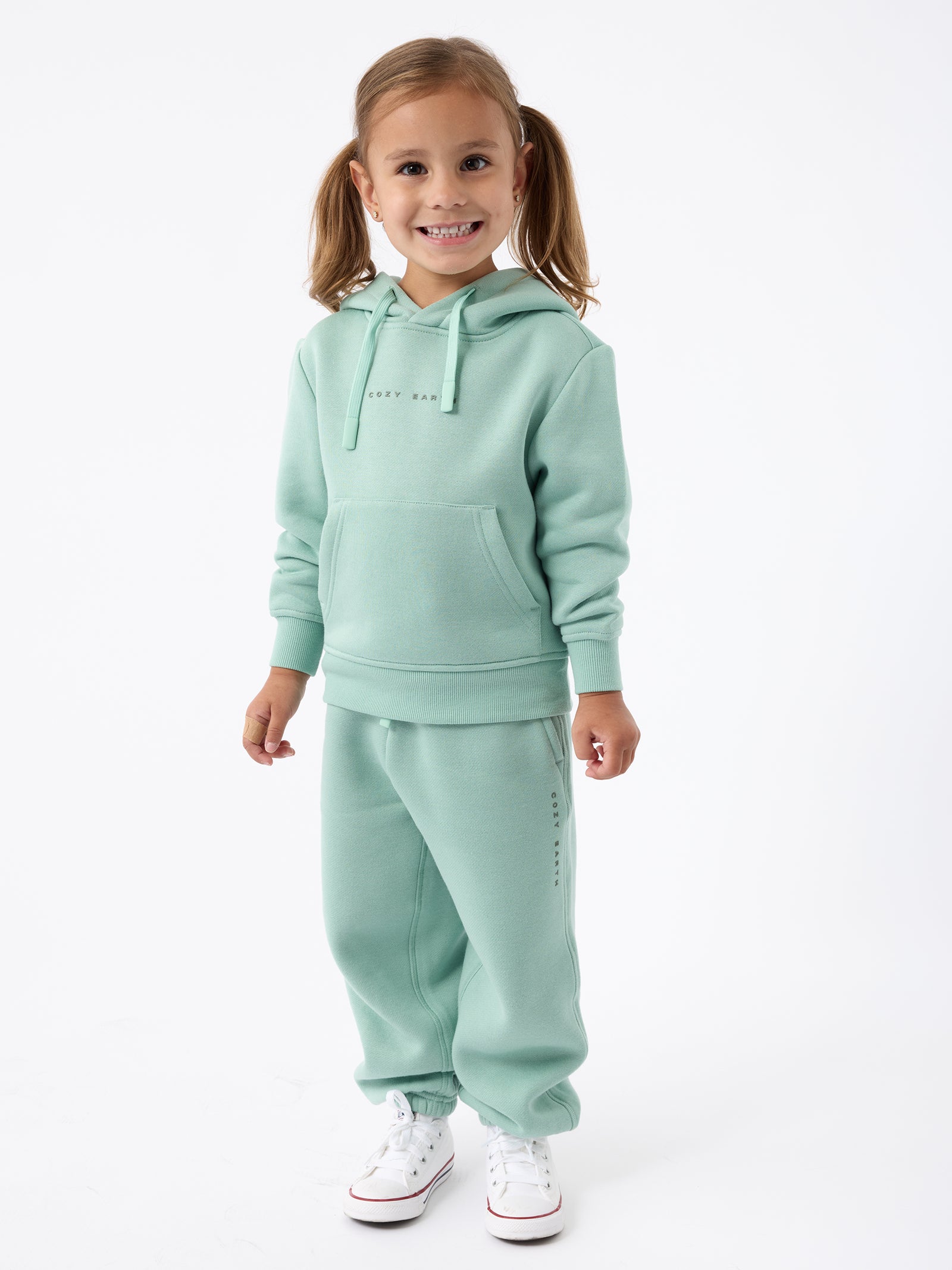 A young girl with light brown hair in pigtails smiles while wearing a Kid's CityScape Hoodie by Cozy Earth along with matching sweatpants. She stands against a white background and is also wearing white sneakers. 