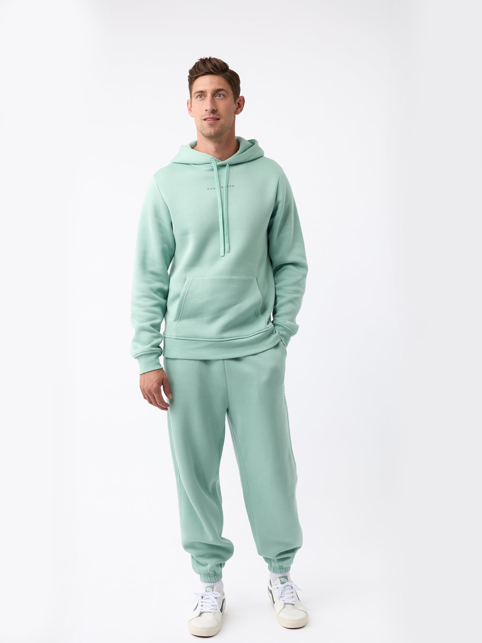 A person is wearing the Men's CityScape Hoodie from Cozy Earth in mint green, paired with matching sweatpants, standing against a plain white background. They complete their outfit with white sneakers featuring green details. 