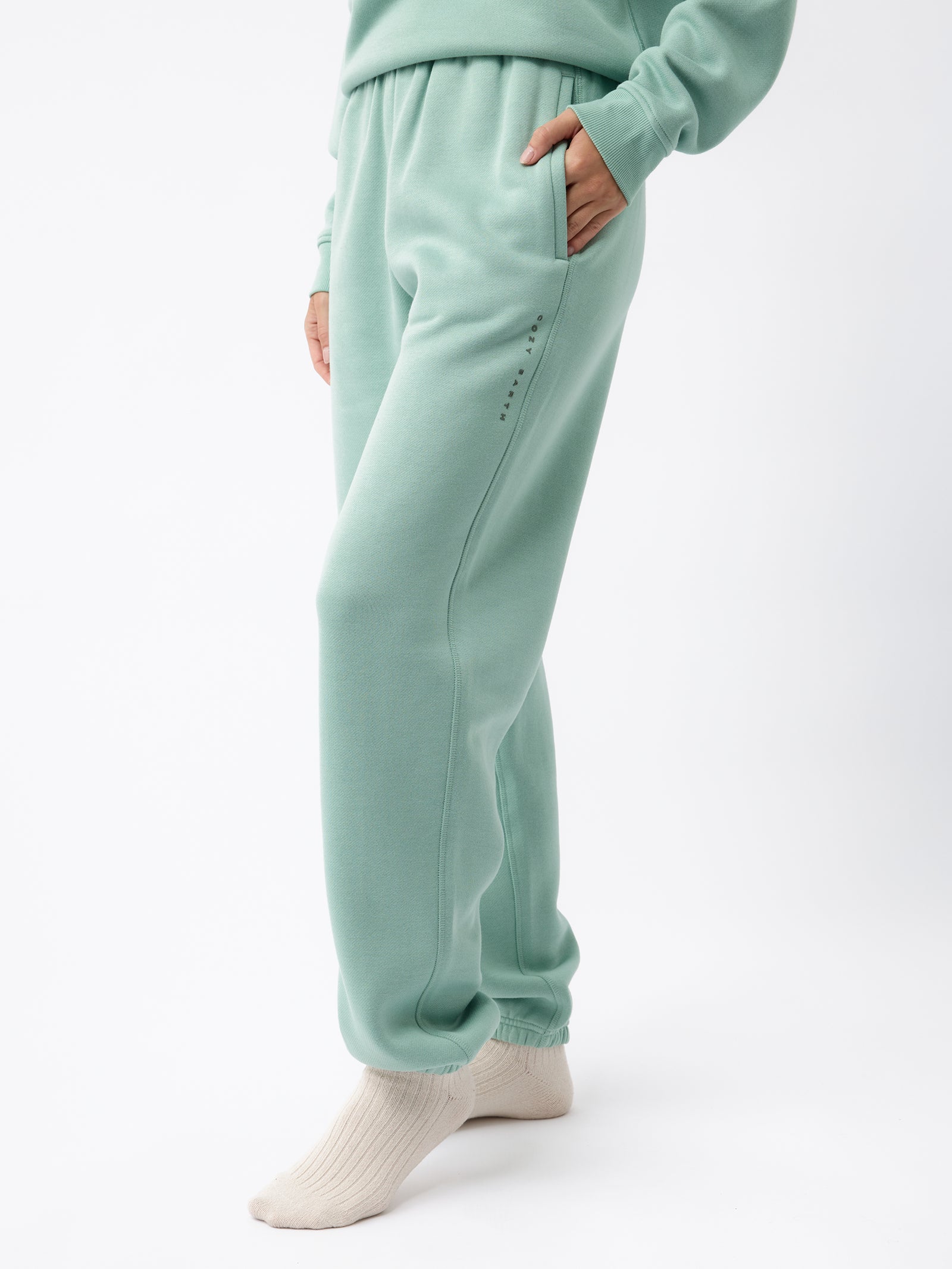 A person wearing Cozy Earth's Women's CityScape Sweatpant in mint green, along with a matching sweatshirt, is standing with one hand in their pocket. They are also wearing cream-colored socks. The background is white, and only the lower half of their body is visible. 