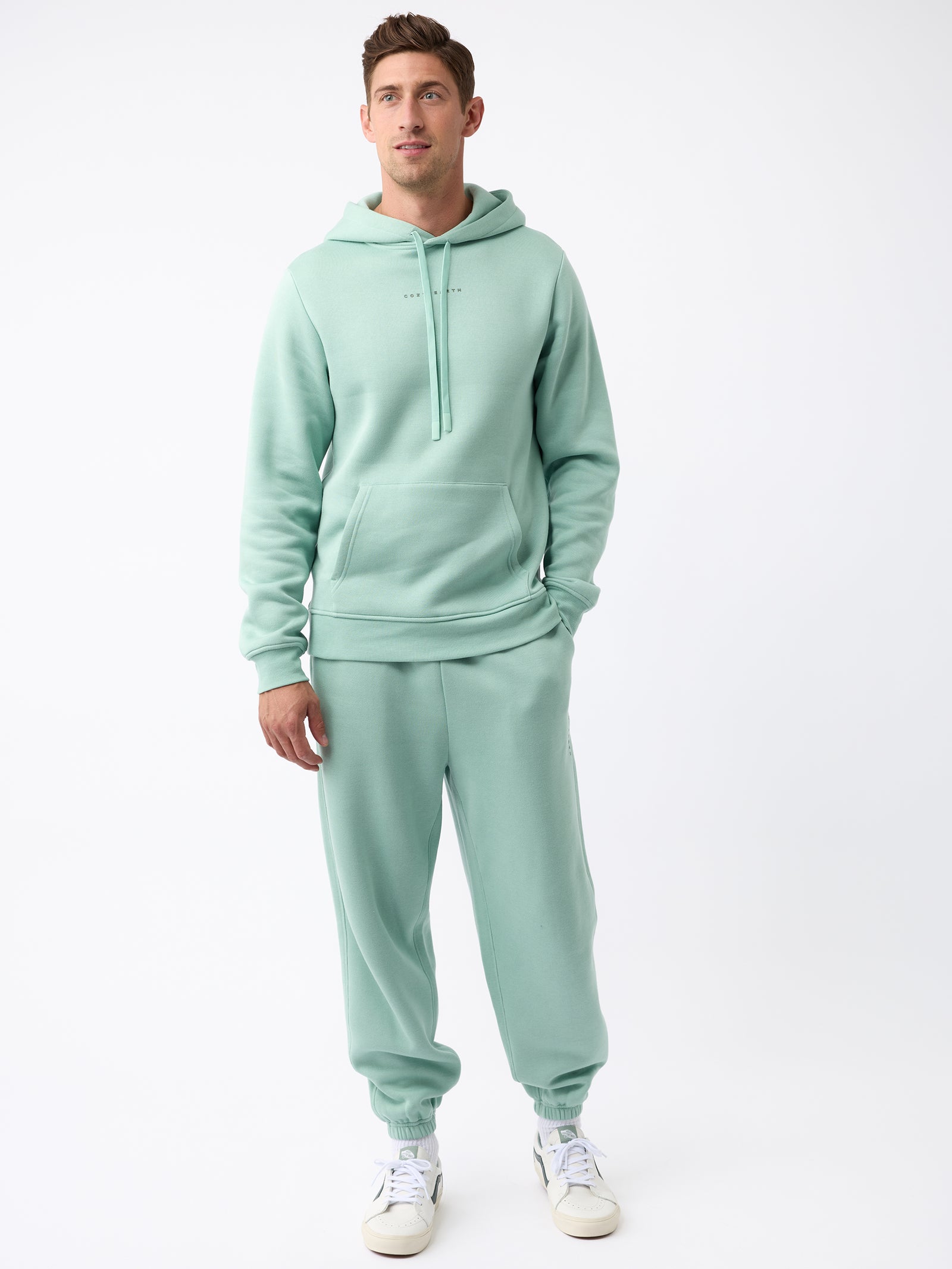 A man dressed in Cozy Earth's Men's CityScape Sweatpant in a light green hue stands against a plain white background. He completes his look with matching attire and white sneakers, his hands resting comfortably at his sides as he gazes slightly to the left. 