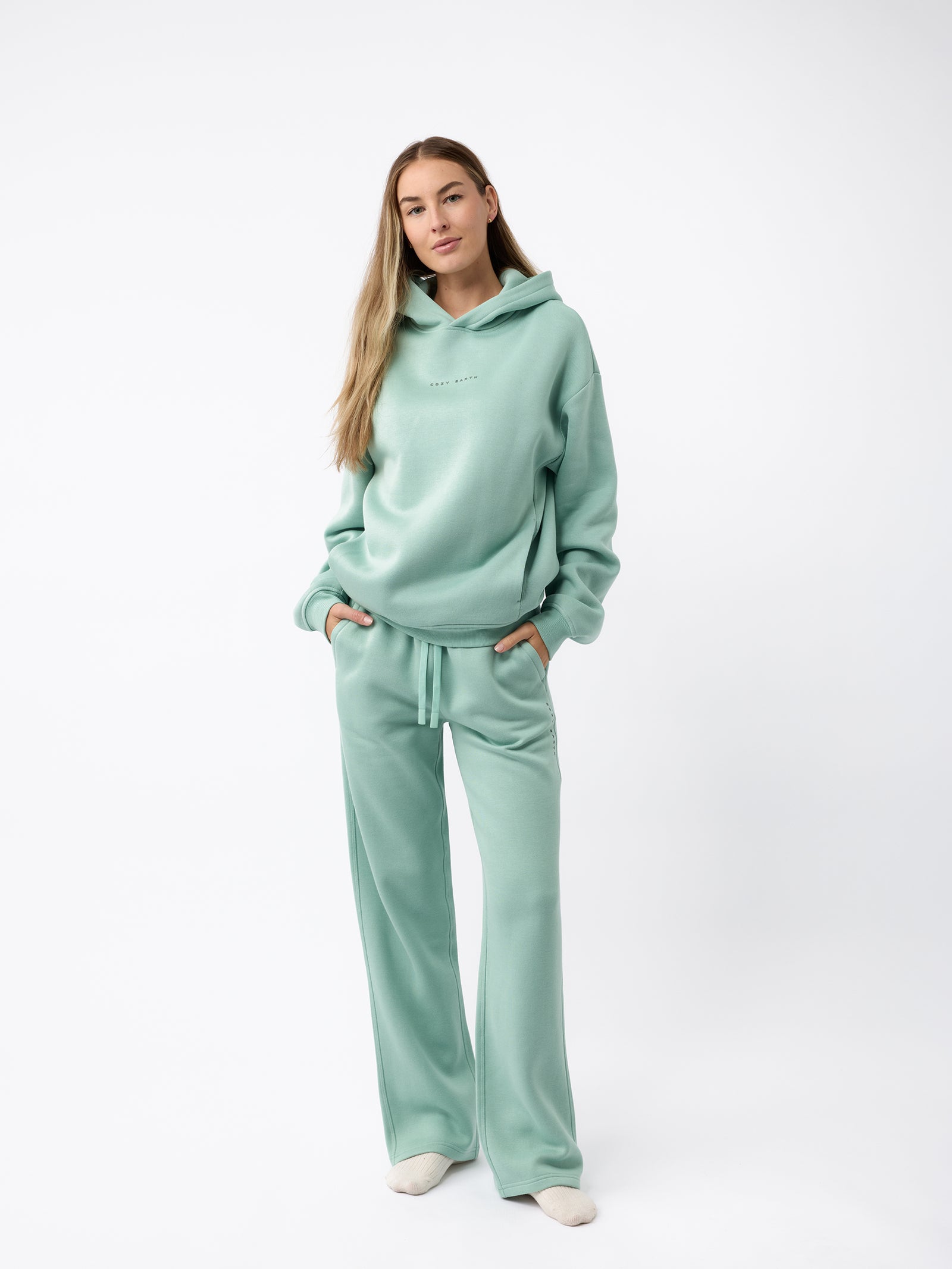 A person stands against a plain backdrop, wearing a coordinated light green hoodie paired with Cozy Earth's Women's CityScape Wide Leg Pant. Their hands rest in the hoodie pockets and their long, light brown hair cascades down, completing a casual and relaxed appearance. 