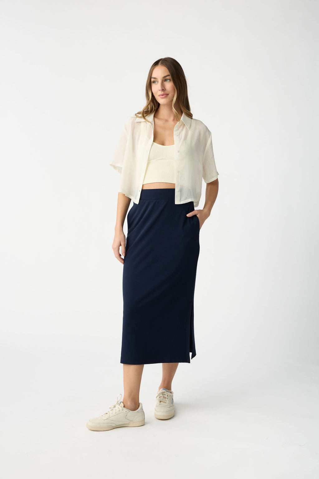 A woman poses casually against a white background, wearing a white short-sleeve button-up shirt over a light top, paired with Cozy Earth's Women's Brushed Bamboo Midi Skirt and white sneakers. One hand rests in her skirt pocket while the other hangs at her side. |Color:Navy