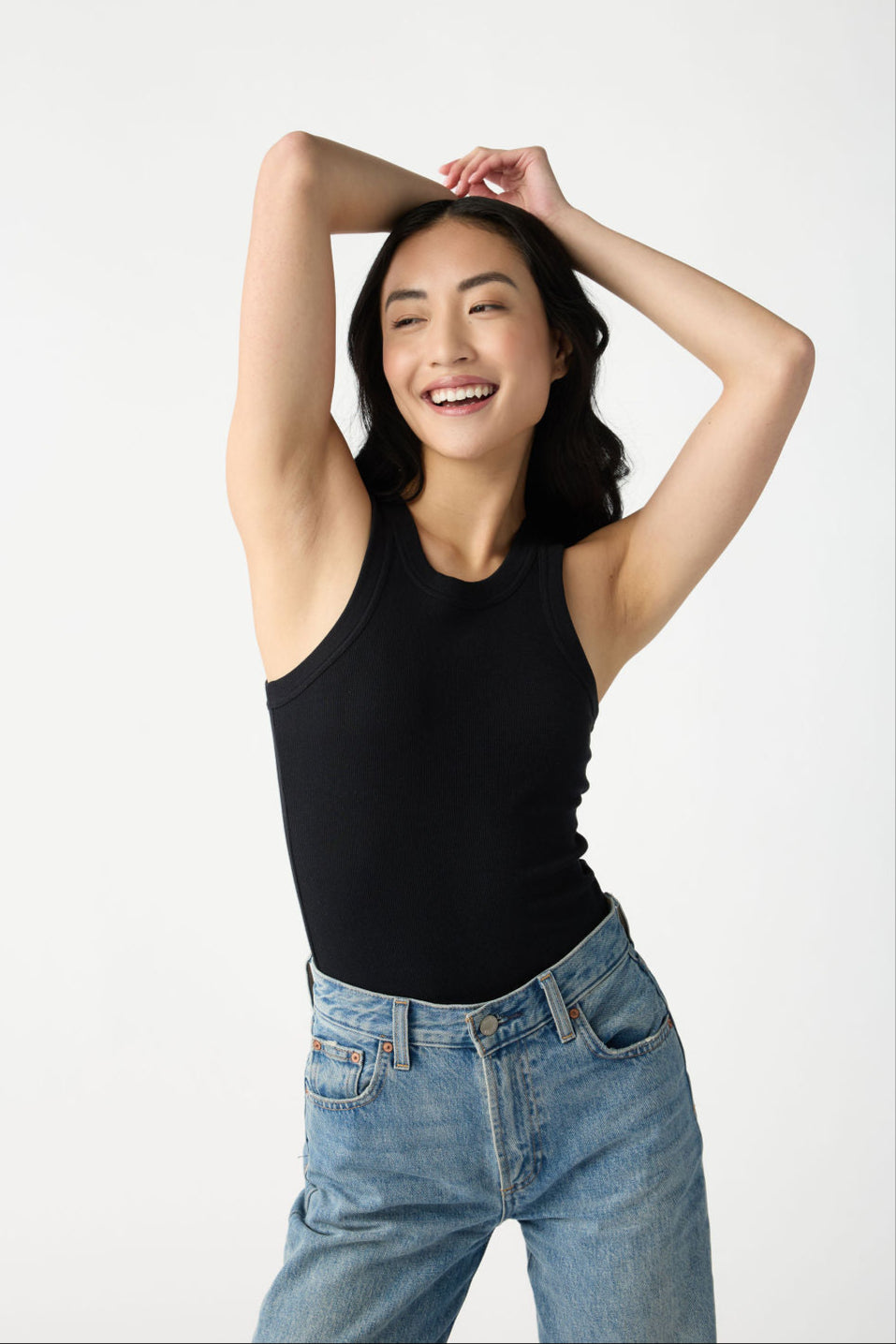 A dark haired woman wears Cozy Earth Women's Fine Ribbed Tank in Jet Black. She rests her arms on her head, smiling, and stands in front of a white background. 