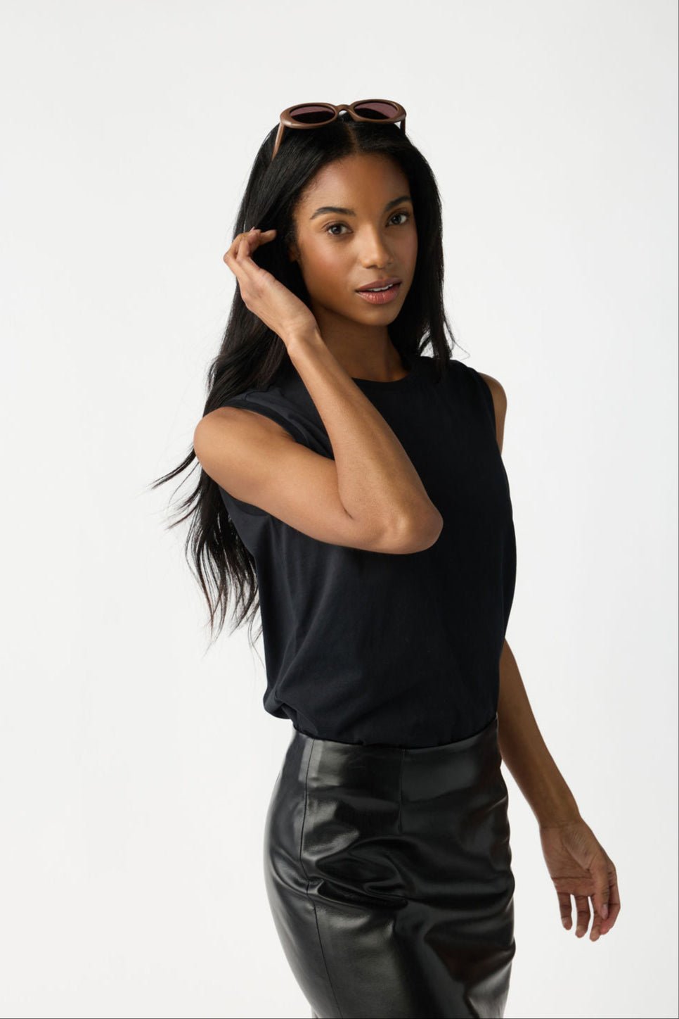 A woman with dark hair and sunglasses on her head wears Cozy Earth Women's All Day Sleeveless Tee in Jet Black. She's tucking her hair behind her ear and is shown from the hips up in front of a white background. |Color:Jet Black