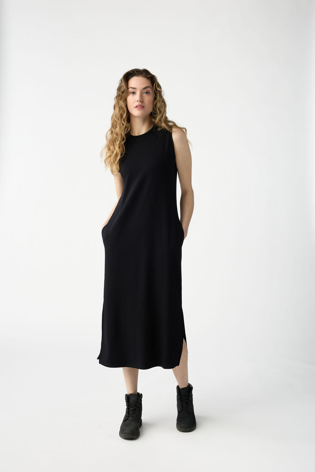 A woman with long curly hair stands against a plain white background, wearing Cozy Earth's Women's Brushed Bamboo Sleeveless Midi Dress and black boots, her hands in the dress pockets. 