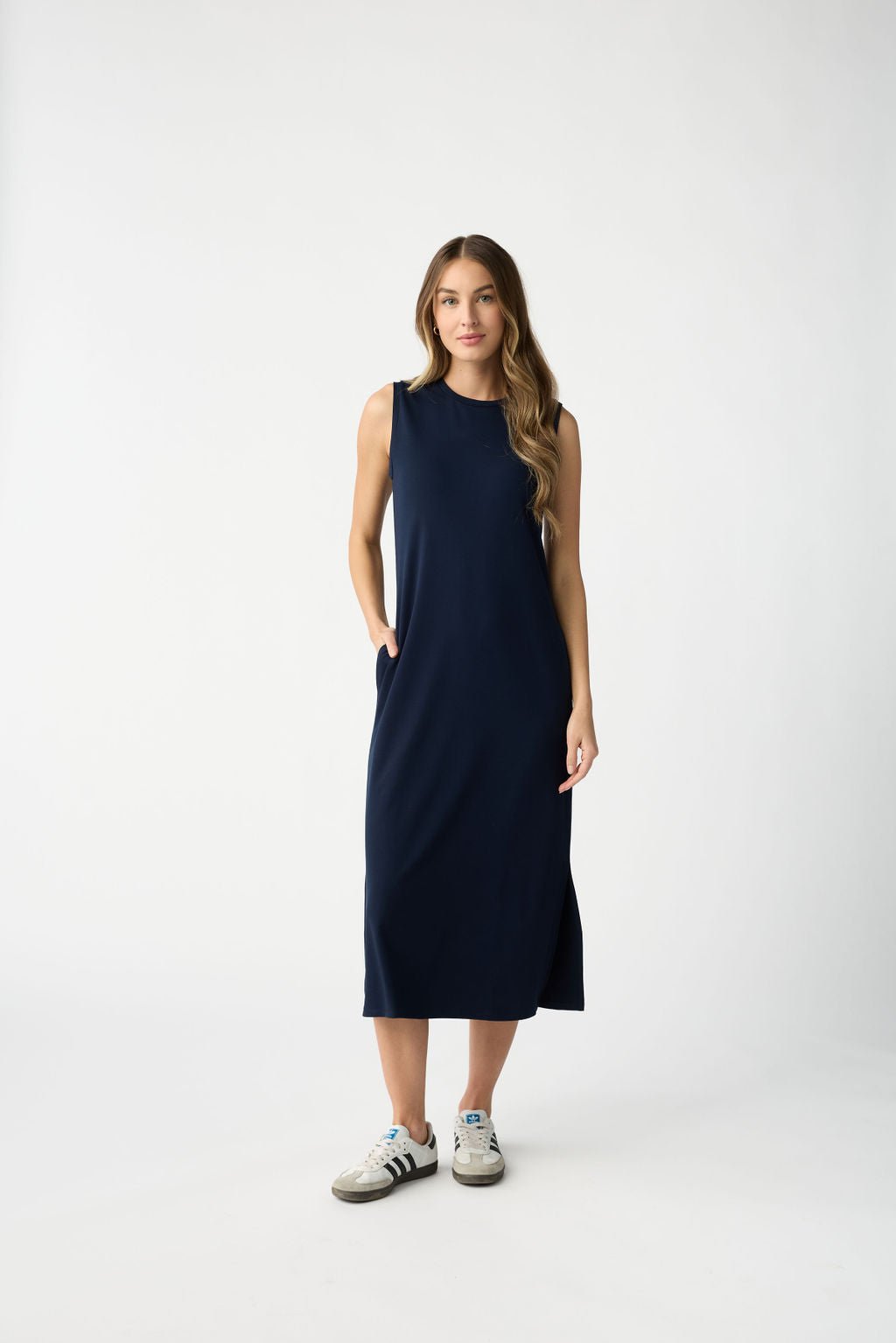 Standing against a plain white backdrop, the person wears Cozy Earth's Women's Brushed Bamboo Sleeveless Midi Dress in dark blue, paired with white sneakers featuring black stripes. Their long, wavy hair cascades down as they place both hands in the dress pockets. 