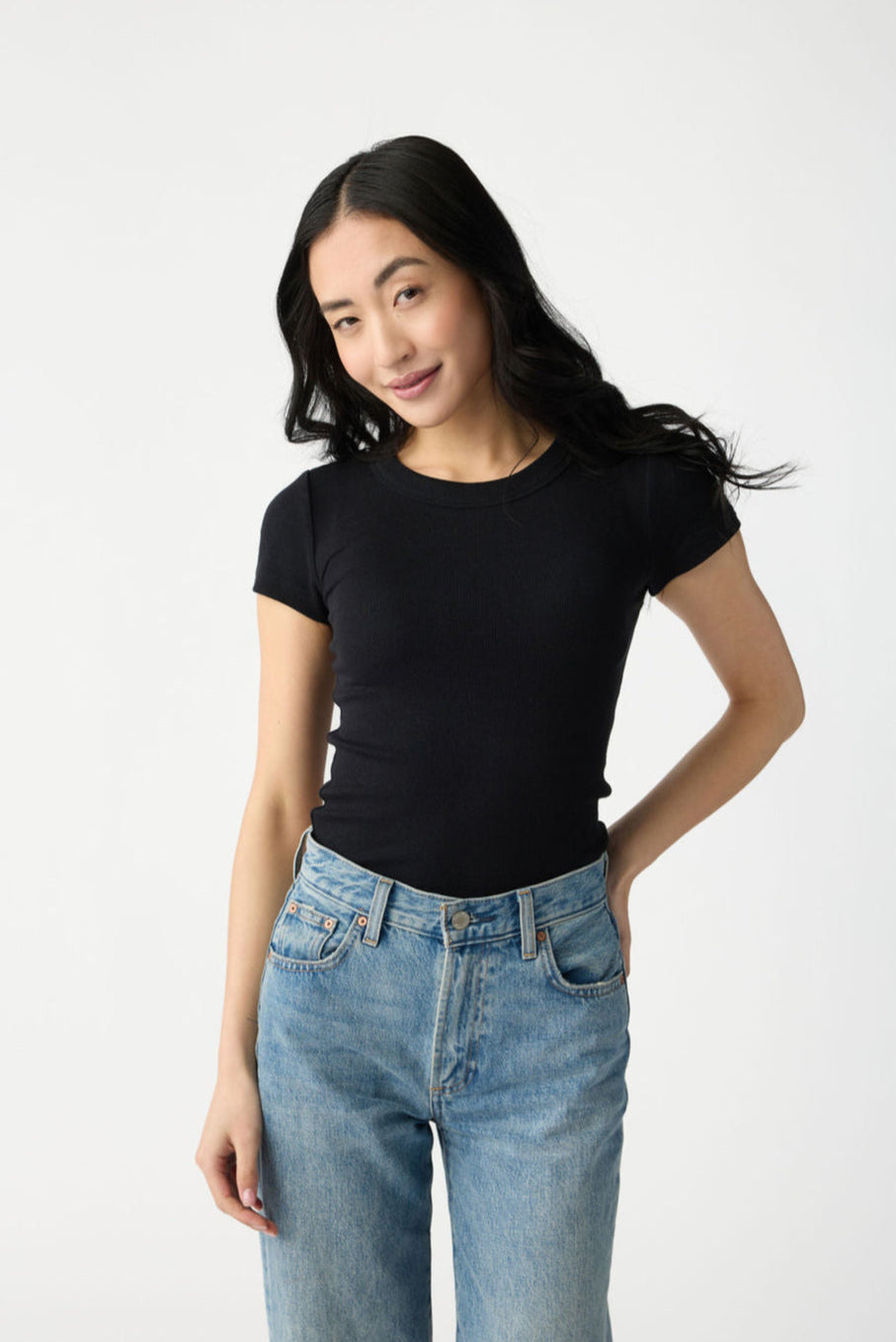 A dark haired woman wears Cozy Earth Women's Fine Ribbed Tee in Jet Black. She is wearing blue jeans and stands in front of a white background. 