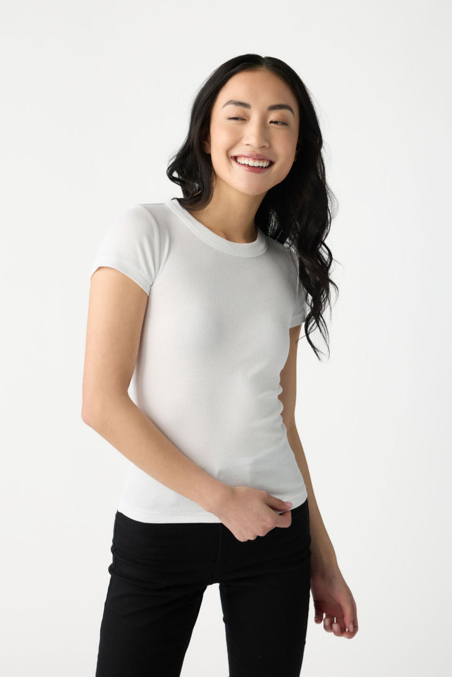 A dark haired woman wears Cozy Earth Women's Fine Ribbed Tee in White. She wears black pants and stands in front of a white background. 