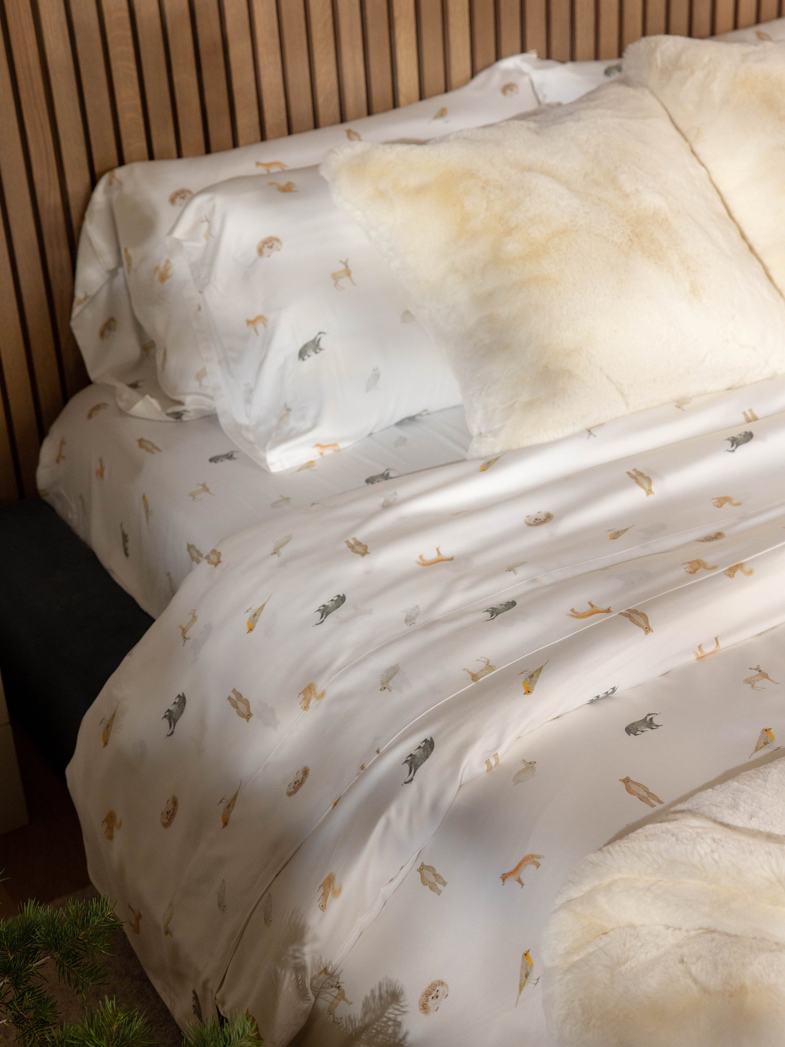 A Bamboo Duvet Cover from Cozy Earth in cream, adorned with small animal prints, complements the cozy bed. The fluffy, light beige pillows add comfort and style against the wooden headboard. In the bottom foreground, a corner of a Christmas tree adds a festive touch to the setting. 