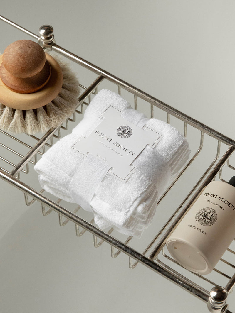 A wire rack holds toiletries: a wooden body brush with bristles on one side, a folded white towel with a "Cozy Earth" label, and a cylindrical container labeled "Complete Cleansing Cloths." The wire rack appears to be part of a shower or bath setup.