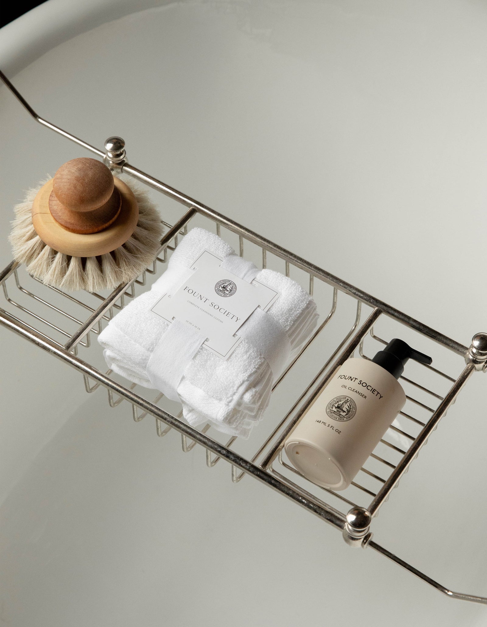 A bathtub tray holds a wooden brush with a rounded handle, a stack of white towels secured with a branded Cozy Earth label, and a beige bottle of bath product labeled "Revitalizing Pair." The background features a clean, white bathtub.