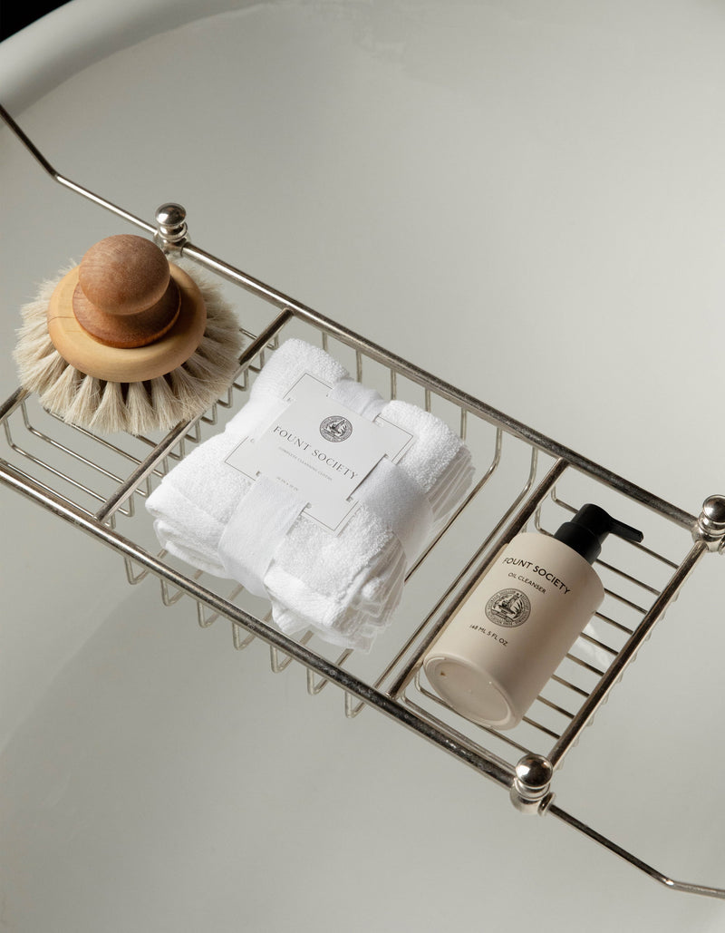 A bathtub tray holds a wooden brush with a rounded handle, a stack of white towels secured with a branded Cozy Earth label, and a beige bottle of bath product labeled "Revitalizing Pair." The background features a clean, white bathtub.