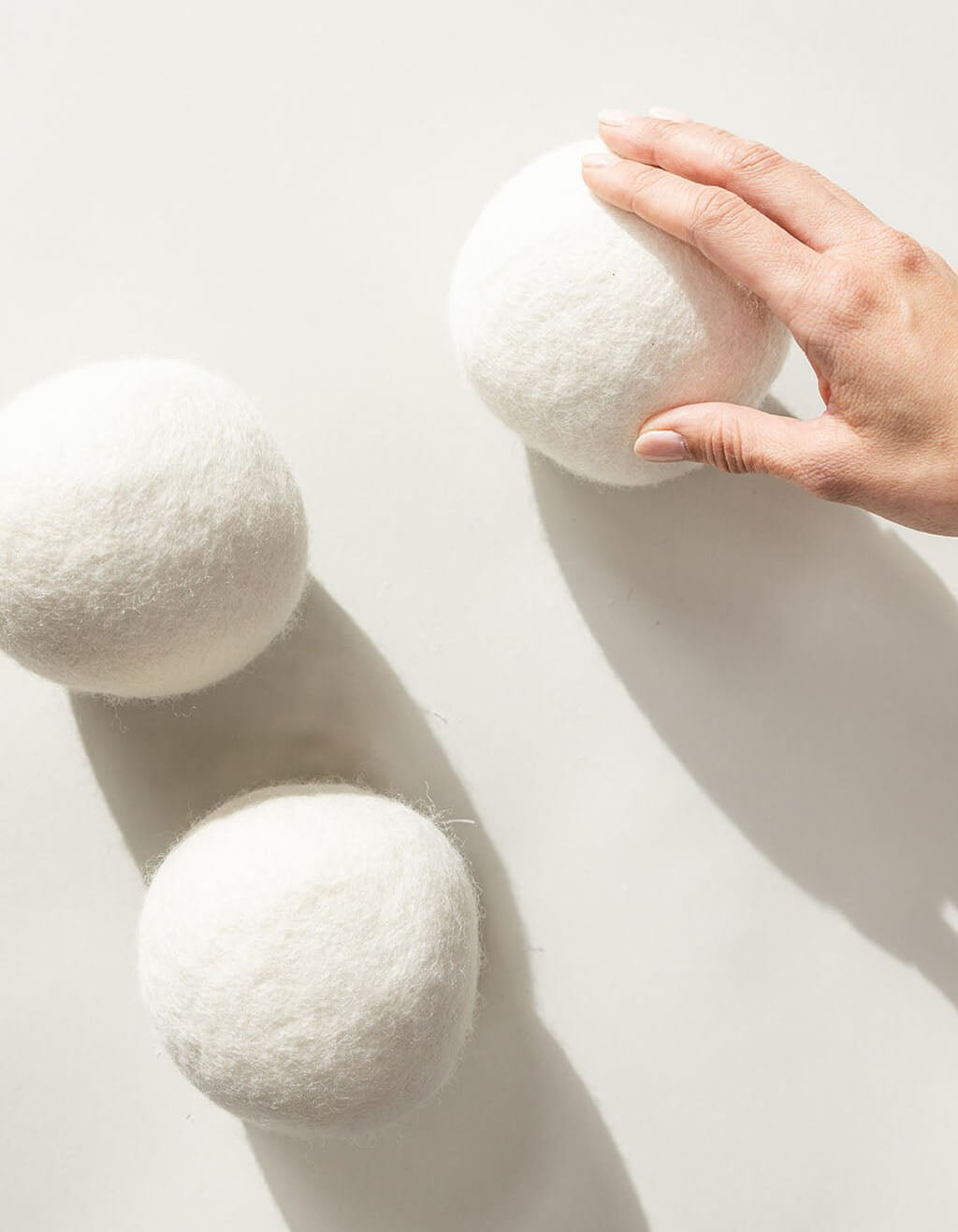 A hand touches one of three large, white Cozy Earth Wool Dryer Balls resting on a light surface, with soft shadows cast beneath them.