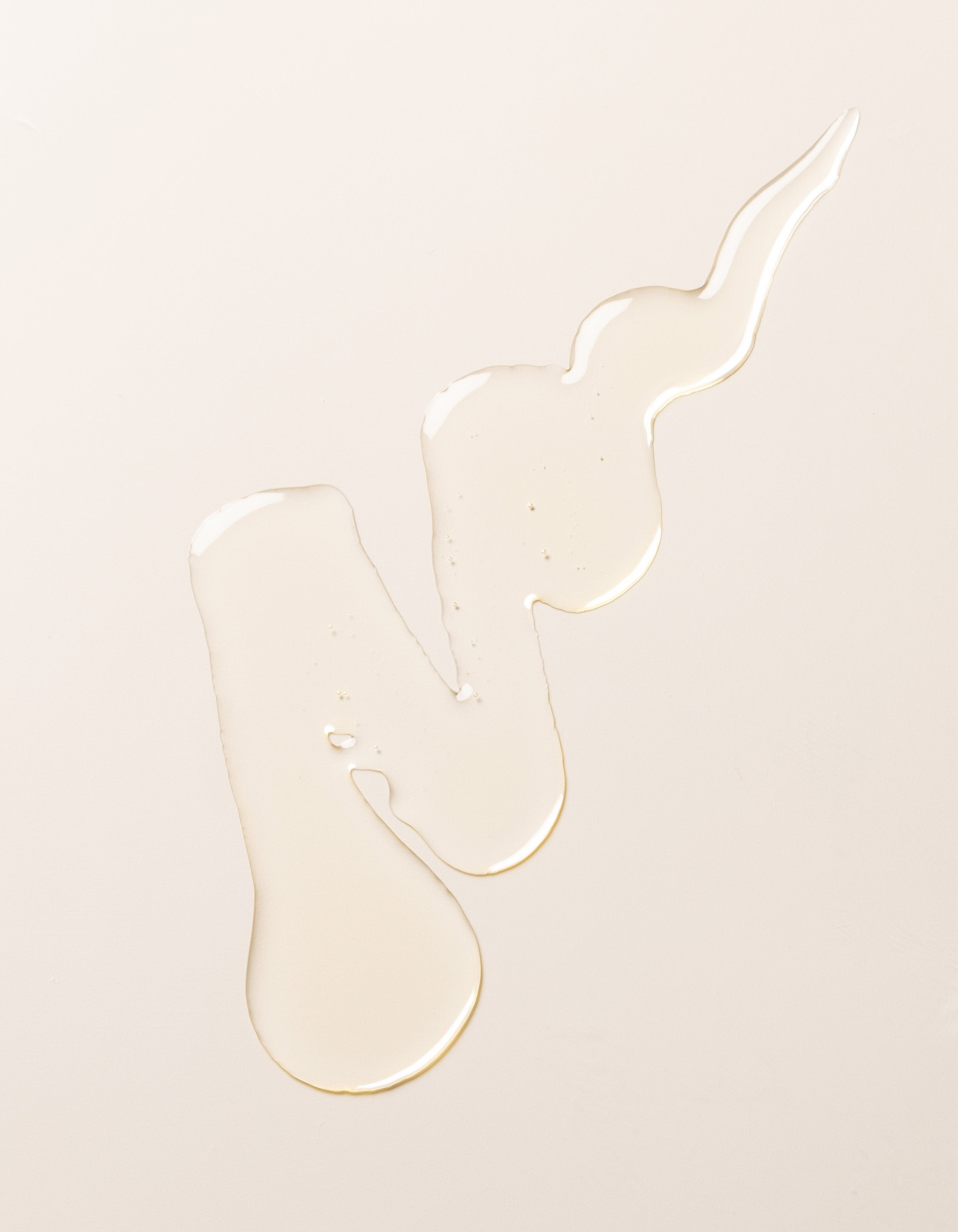 A swirl of transparent, light-colored Cozy Earth's Revitalizing Pair liquid on a plain, pale beige surface. The liquid forms irregular shapes with droplets at the ends, creating a visually interesting pattern.
