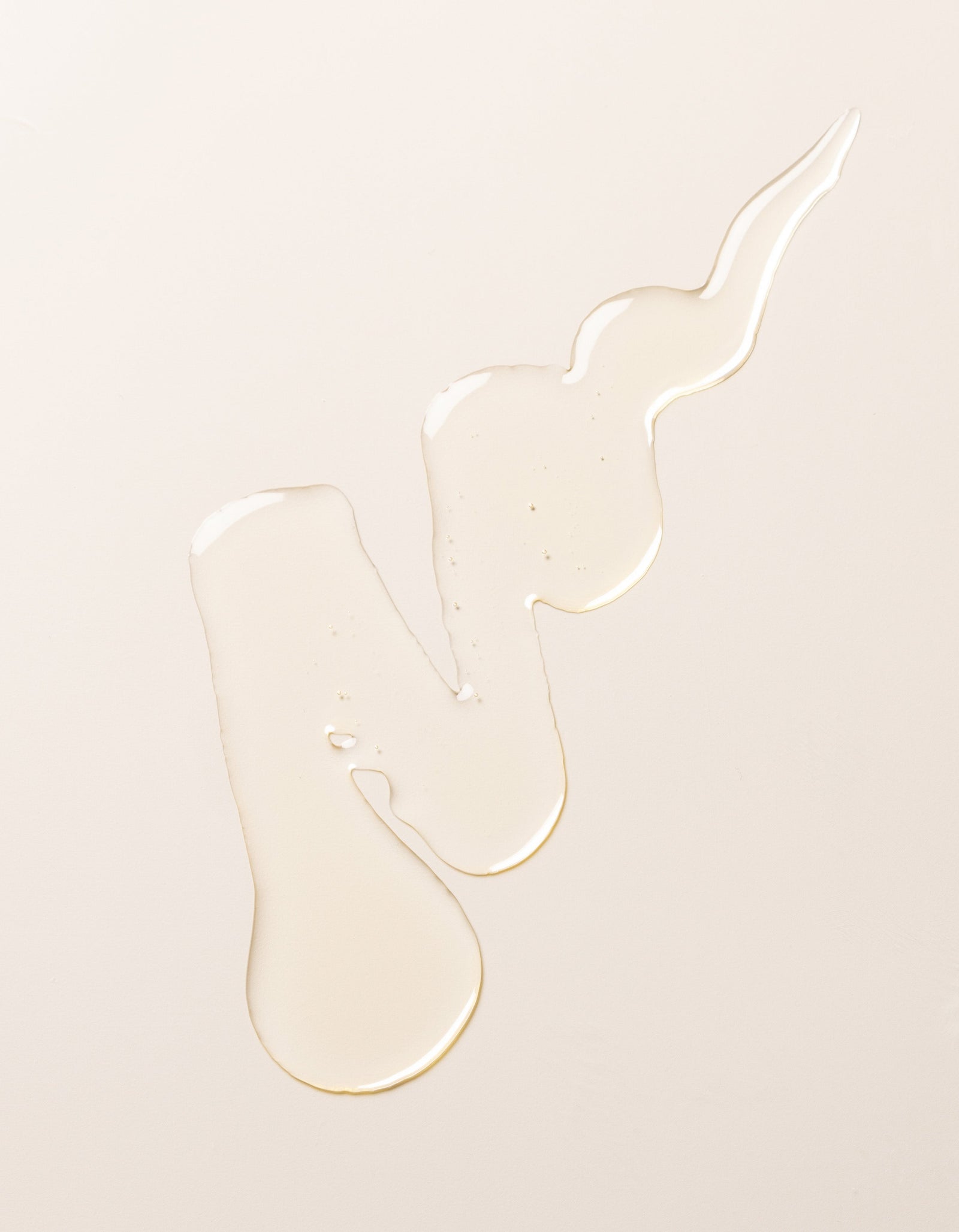A swirl of transparent, light-colored Cozy Earth's Revitalizing Pair liquid on a plain, pale beige surface. The liquid forms irregular shapes with droplets at the ends, creating a visually interesting pattern.