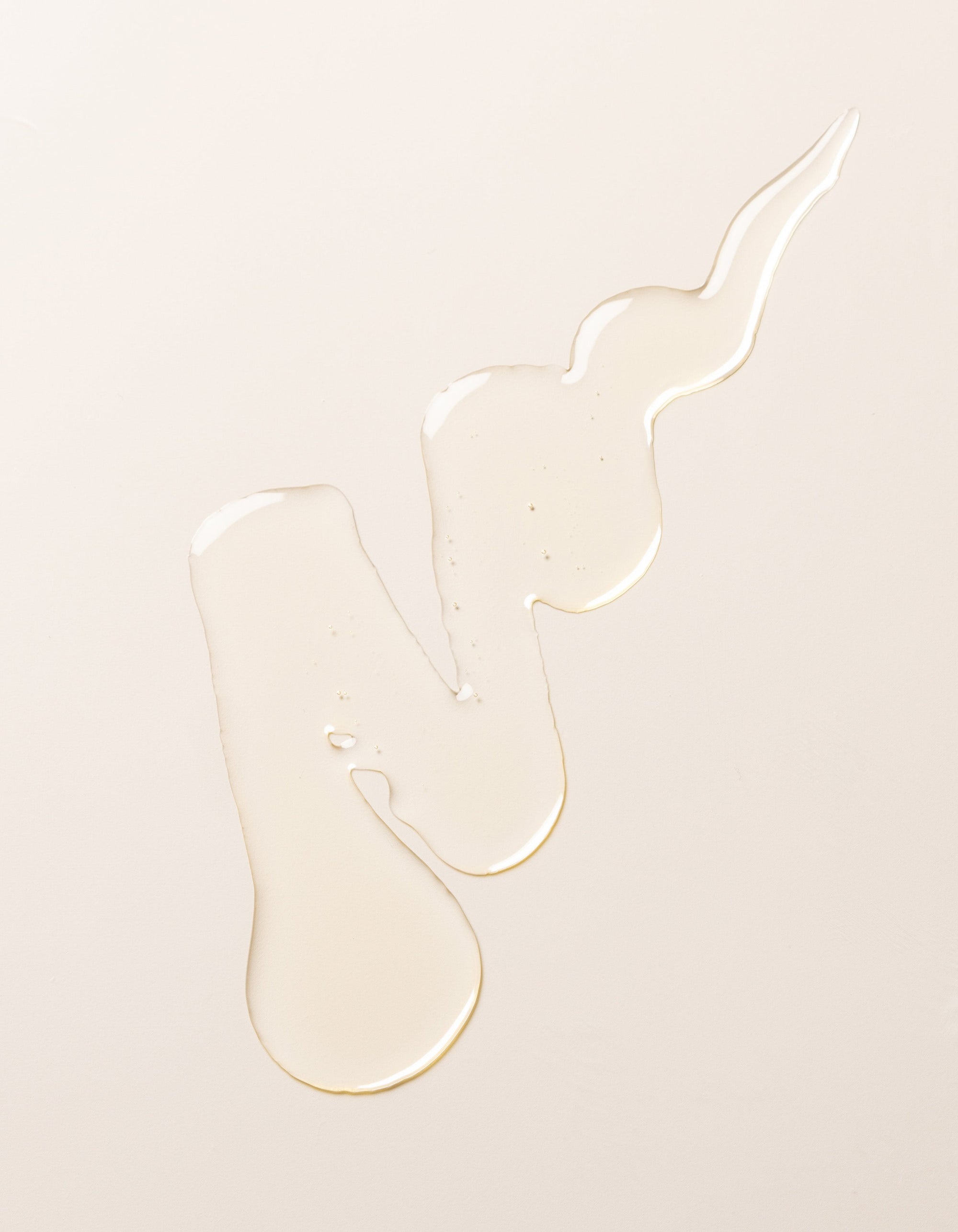 A swirl of transparent, light-colored Cozy Earth's Revitalizing Pair liquid on a plain, pale beige surface. The liquid forms irregular shapes with droplets at the ends, creating a visually interesting pattern.