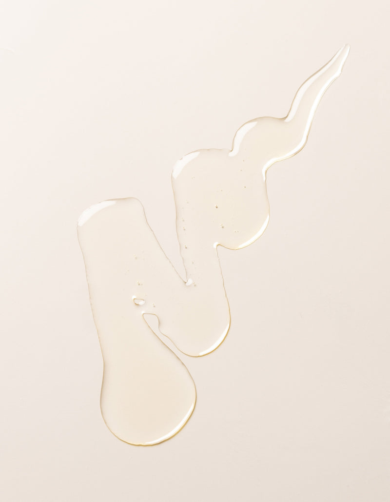 An irregular-shaped spill of Cozy Earth's Oil Cleanser on a smooth, light-colored surface. The clear liquid forms a long, winding pattern with droplets and uneven edges, creating the impression of fluid motion.