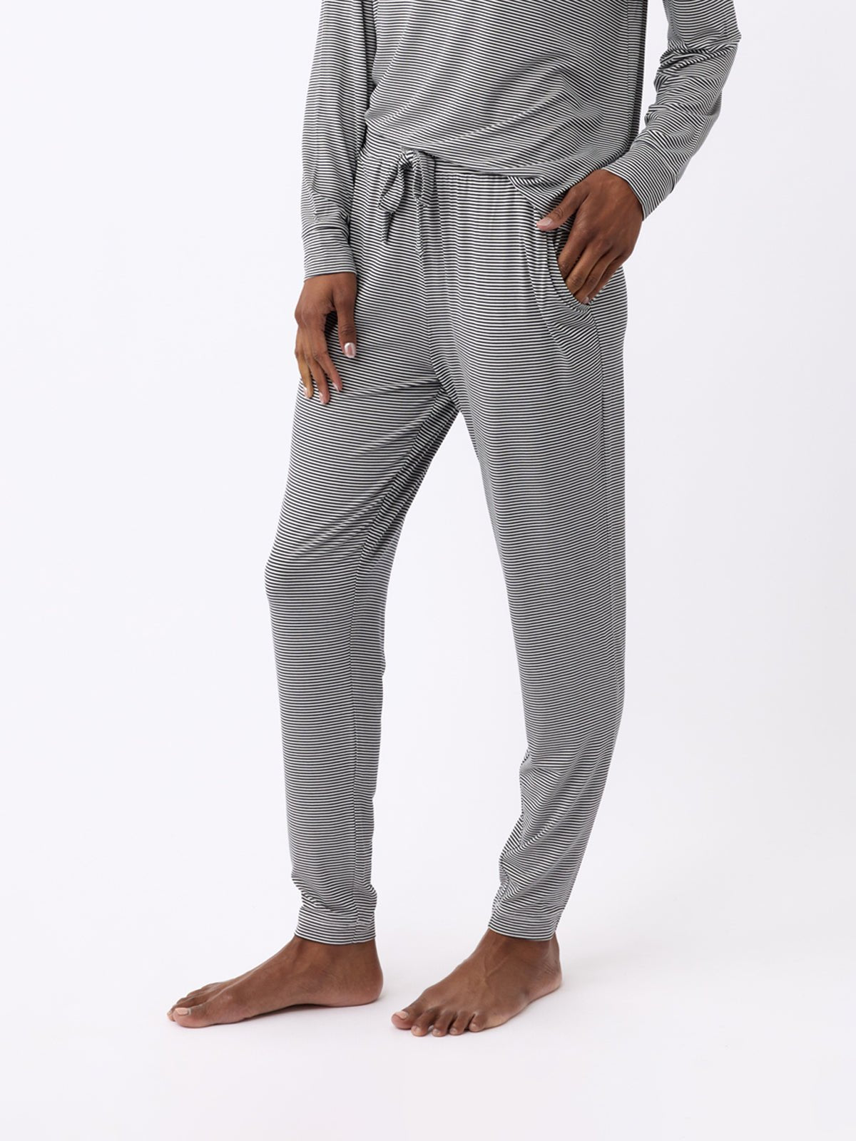 A person models Cozy Earth's Women's Bamboo Stretch-Knit Tapered Pants, featuring gray stripes and a matching long-sleeved top. The photo highlights the pants' lower half against a plain white background, with one hand in a pocket. 