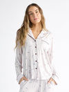 A person wearing Cozy Earth Women's Bamboo Stretch-Knit Long Sleeve Pajama Top, featuring a collar, buttons, and chest pocket, stands relaxed against a white background with long hair and hands in pockets. The light-colored top has a subtle print. |Color:French Lavender
