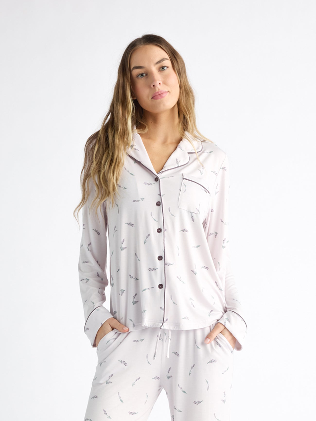 A person wearing Cozy Earth Women's Bamboo Stretch-Knit Long Sleeve Pajama Top, featuring a collar, buttons, and chest pocket, stands relaxed against a white background with long hair and hands in pockets. The light-colored top has a subtle print. 