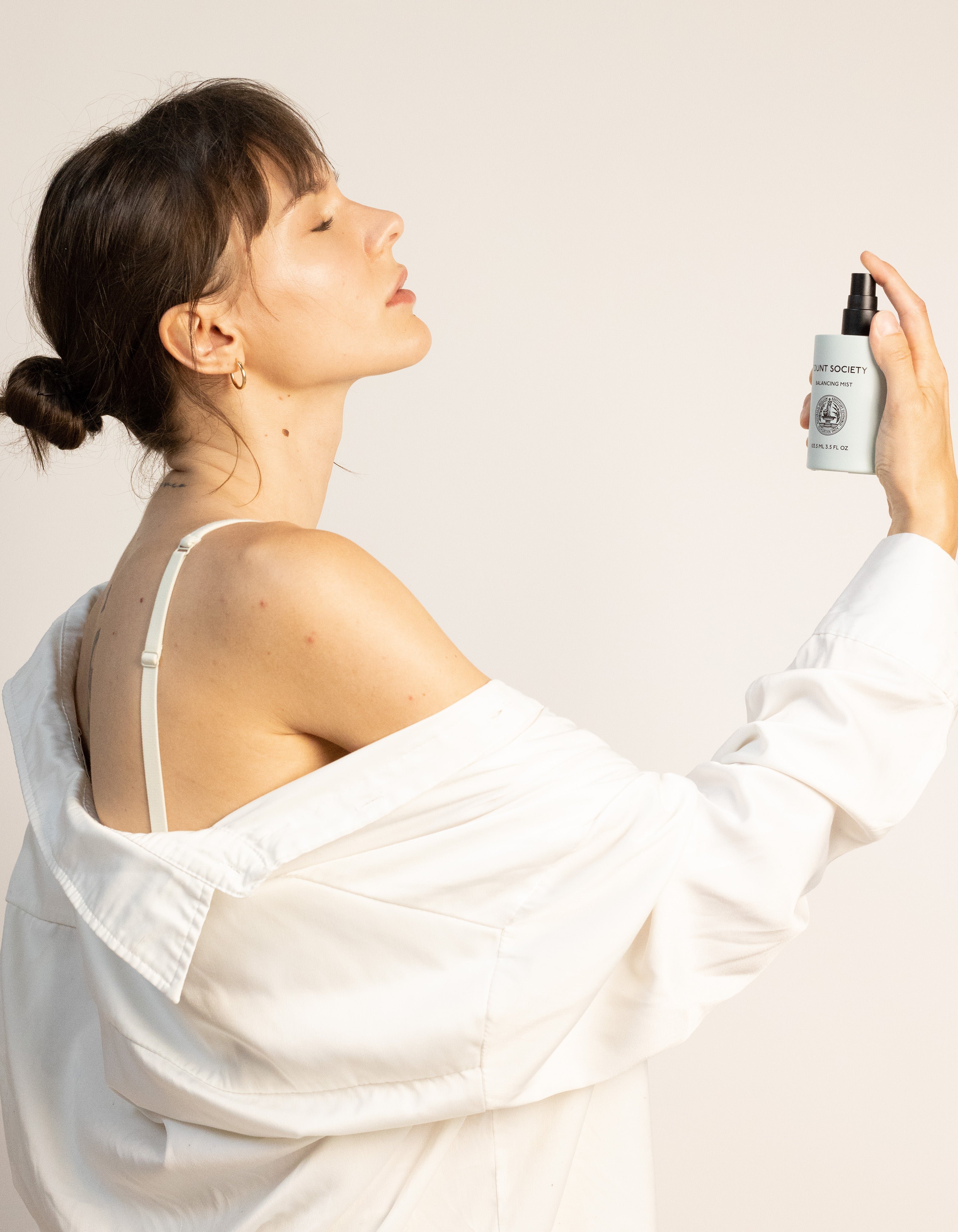 A woman with dark hair tied in a bun holds a bottle of Cozy Earth's Balancing Face Mist near her face, spraying it with a relaxed expression. She is wearing a white shirt that slips off one shoulder, revealing a white strap underneath. The background is plain and light-colored. 