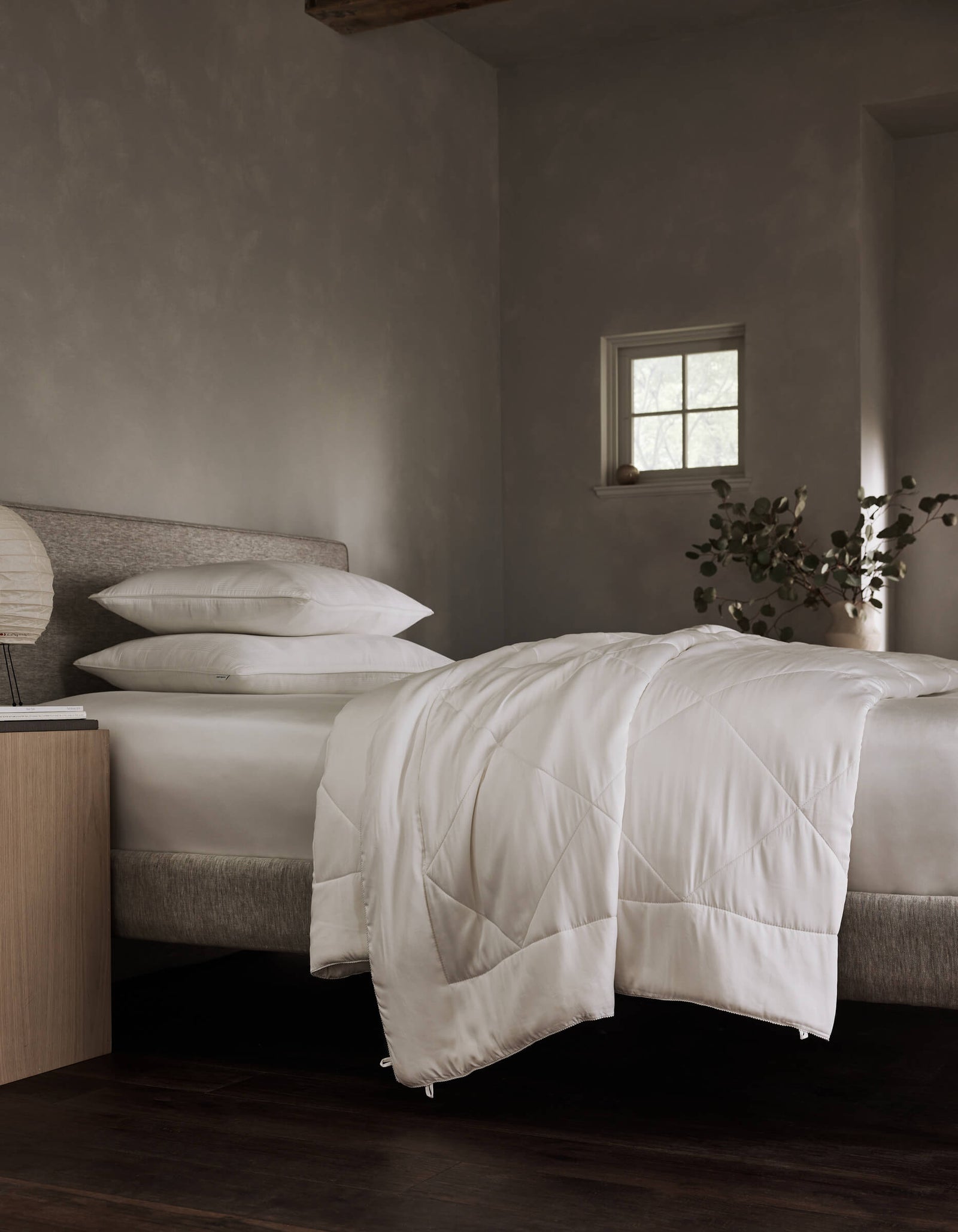 A neatly made bed features Cozy Earth's Bamboo Insert Bundle bedding and pillows in a softly lit, minimalist bedroom with gray walls, a small window, a corner plant, and a wooden nightstand holding a lamp and book.