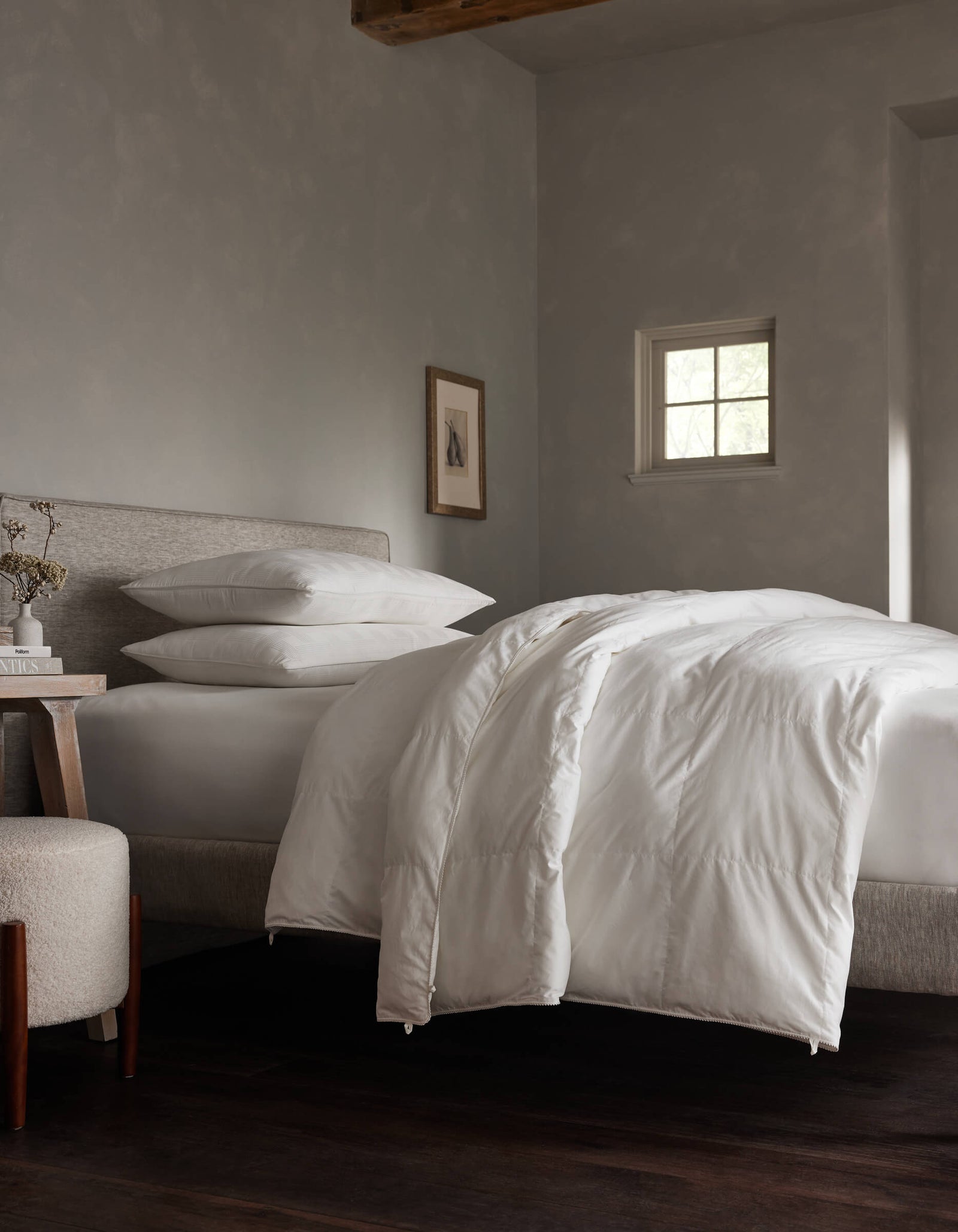 A neatly made bed features the Soft-Wash Cotton Bedding Deluxe Bundle - Down Alternative by Cozy Earth, with white bedding and pillows in a softly lit, minimalist bedroom, accented by neutral walls, framed art, and a bedside table with books and a vase.