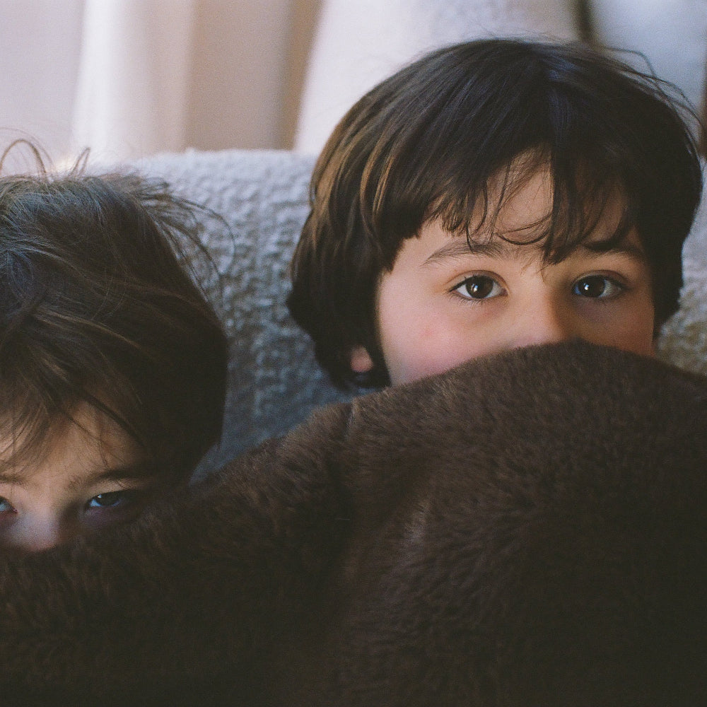 Two children peeking from behind a blanket with a neutral background