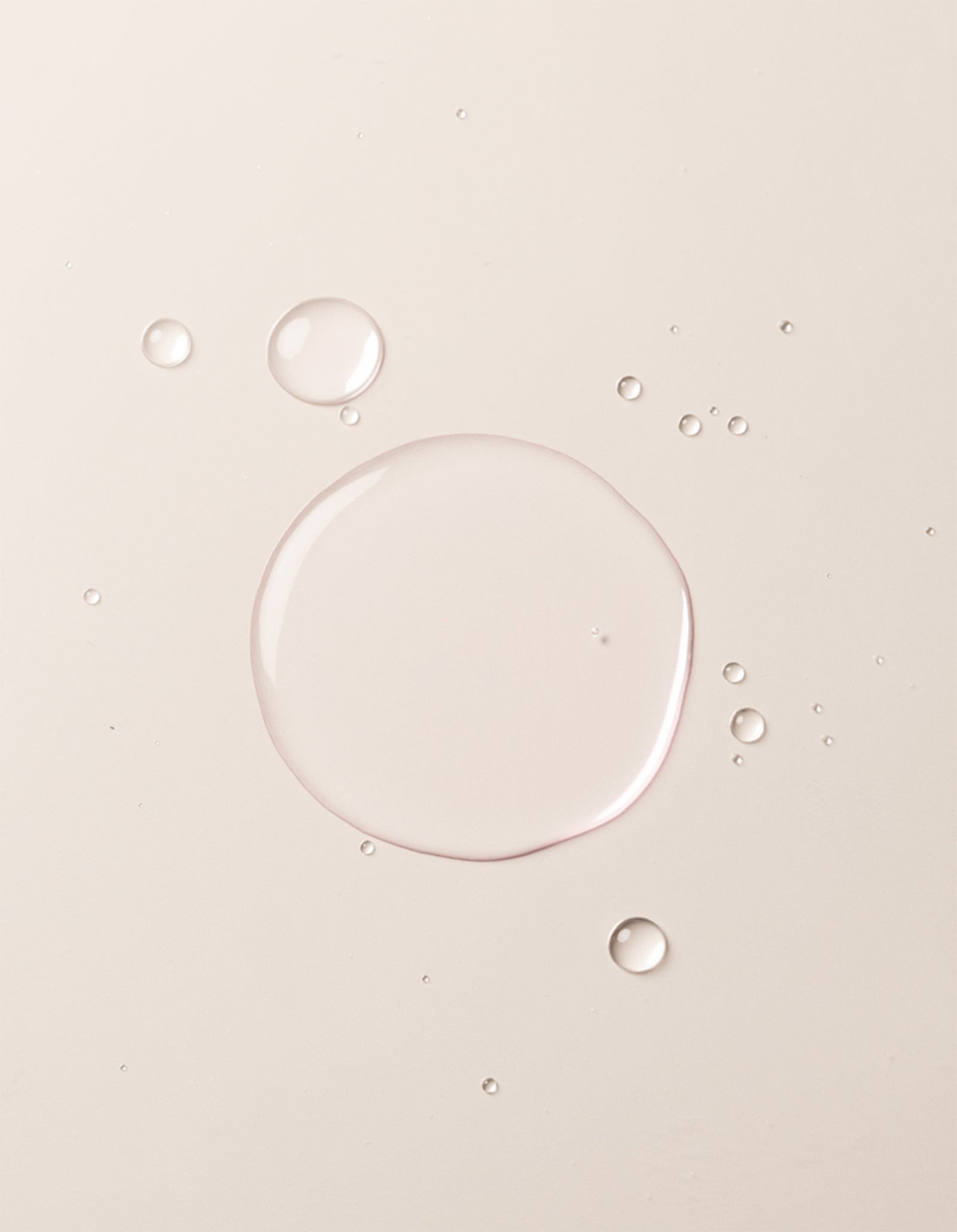 Clear liquid droplets of Balancing Face Mist by Cozy Earth are scattered on a smooth, light beige surface. The largest droplet is centered, surrounded by smaller drops, giving the image a minimal, clean, and soft appearance. 