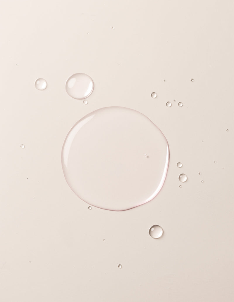 A close-up image showcases translucent liquid droplets of Cozy Earth's Balancing Face Mist on a smooth, off-white surface. The droplets vary in size, with one large central droplet surrounded by several smaller ones, creating a clean and minimalistic appearance.