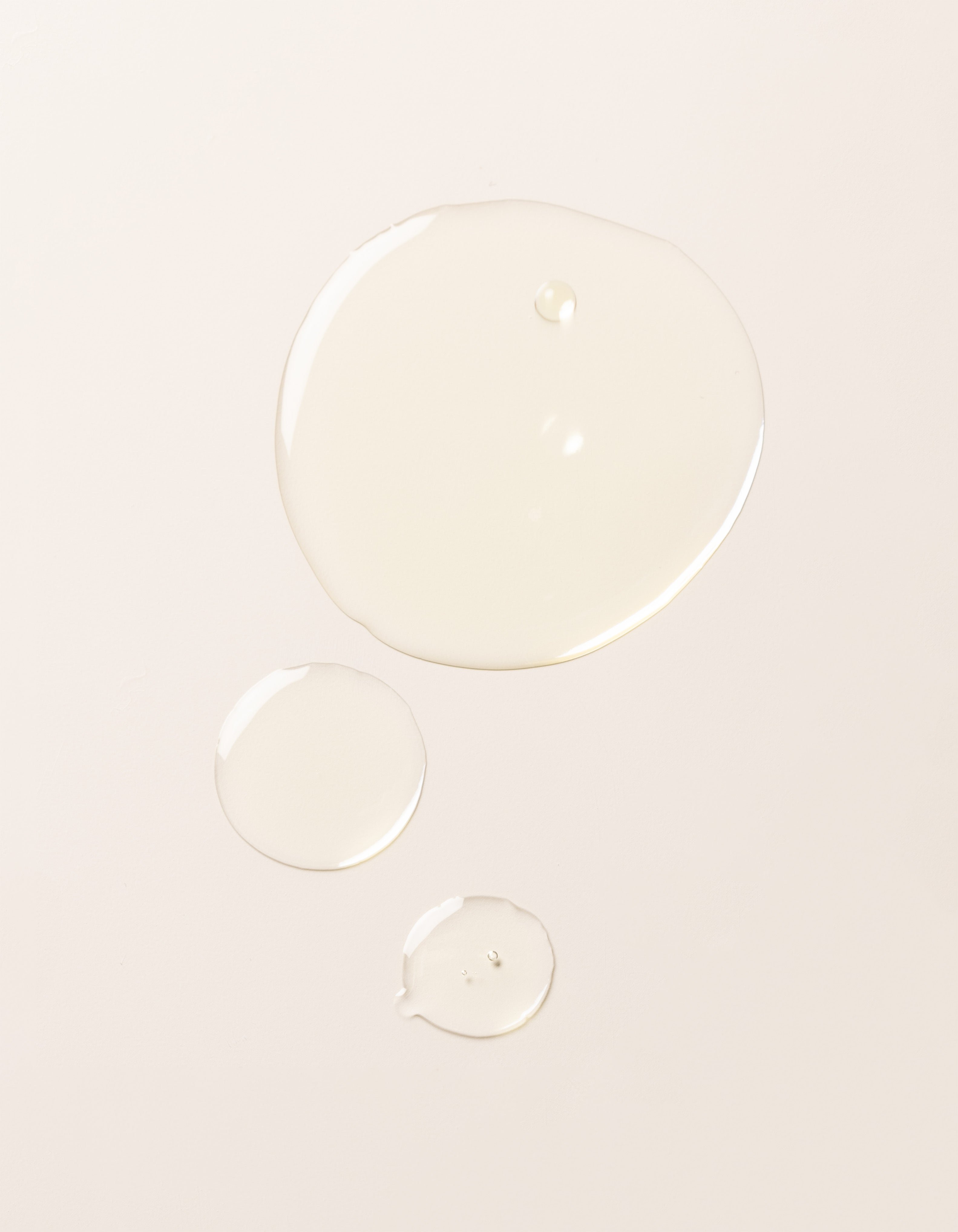 Three droplets of Cozy Earth's Brightening Vitamin C Oil, each of a different size, are spaced apart on a smooth, light-colored surface.