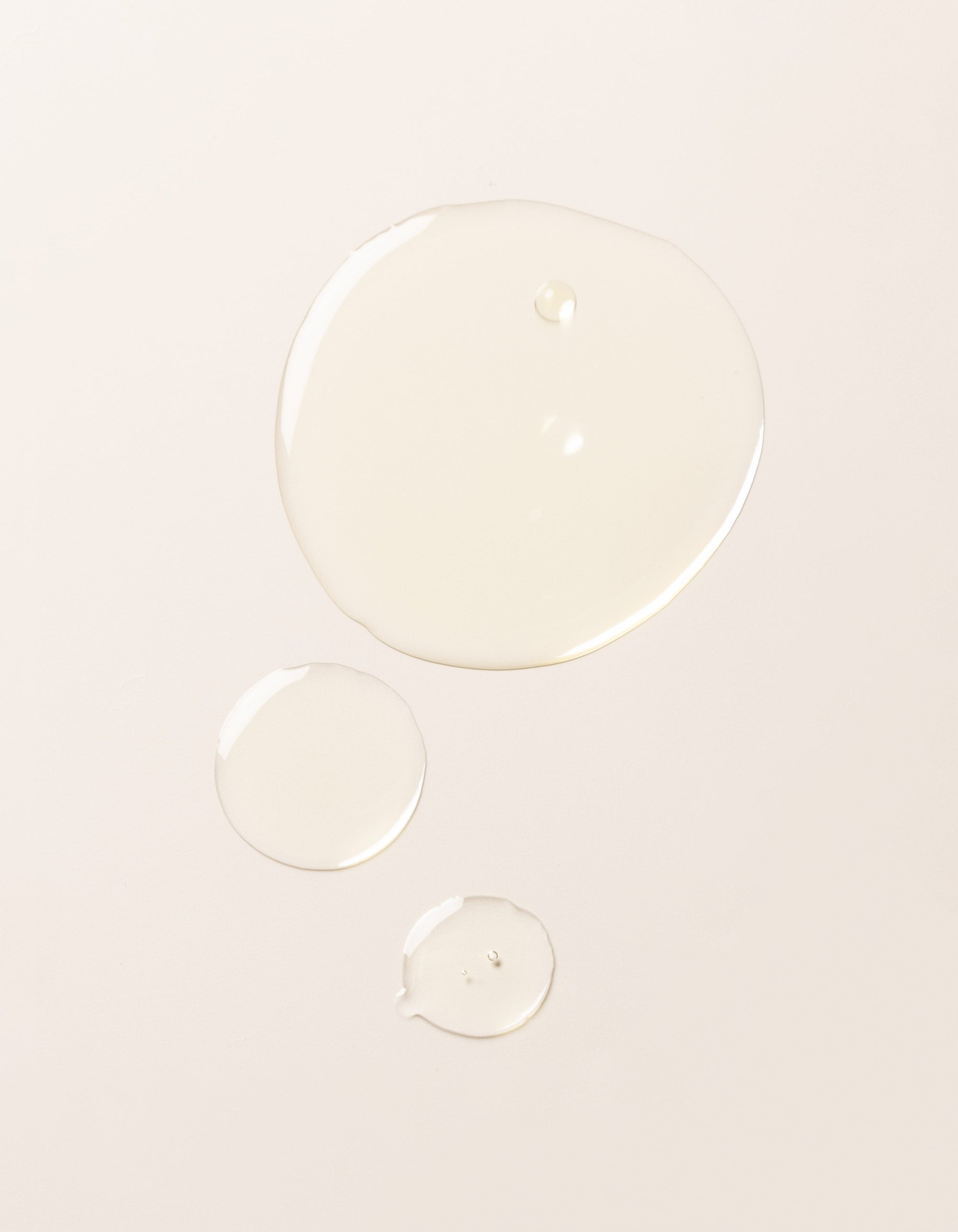 Three droplets of Cozy Earth's Brightening Vitamin C Oil, each of a different size, are spaced apart on a smooth, light-colored surface.