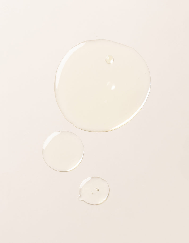 Three droplets of Cozy Earth's Brightening Vitamin C Oil, each of a different size, are spaced apart on a smooth, light-colored surface.