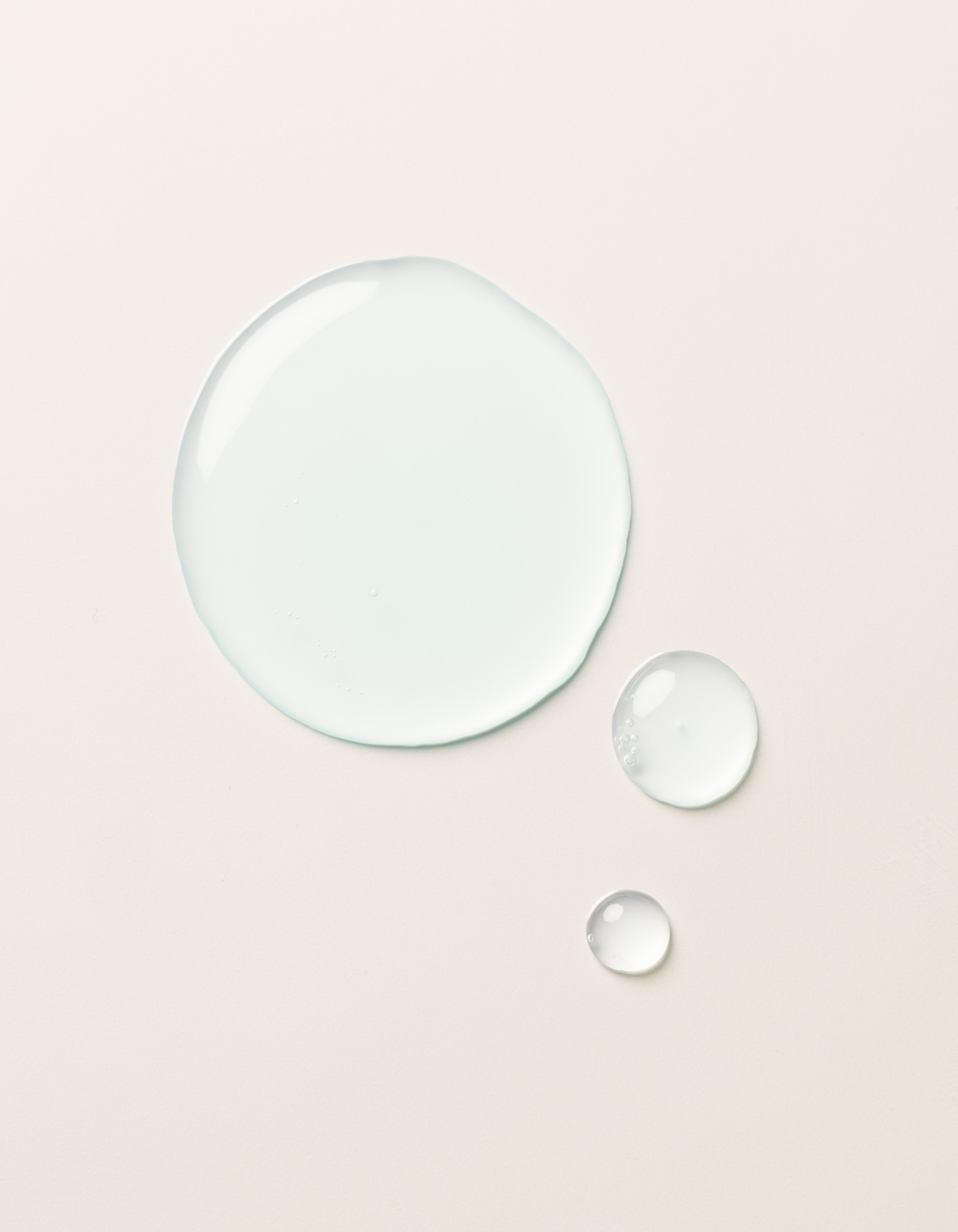 A large clear droplet and two smaller droplets of Cozy Earth Hydrating Serum rest on a smooth, light beige surface. The glossy, semi-transparent droplets reflect light slightly on their surfaces.