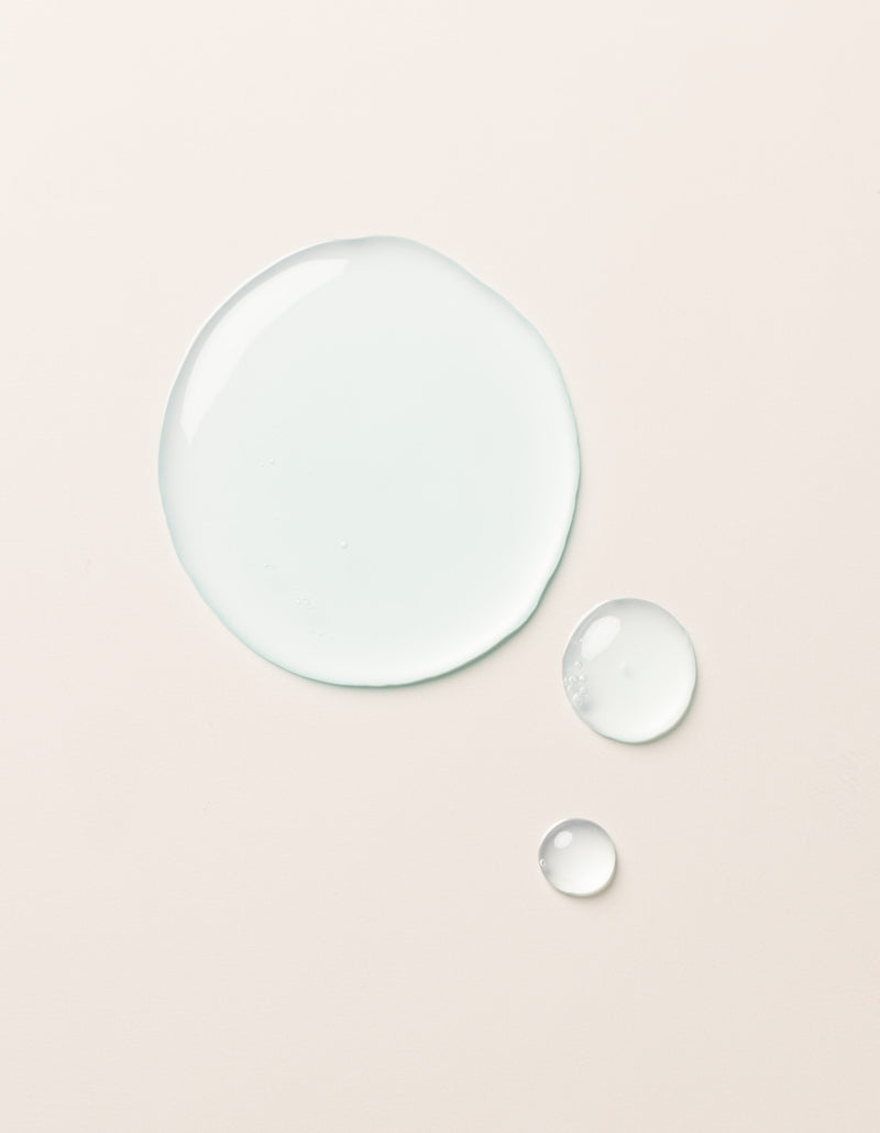 A large clear droplet and two smaller droplets of Cozy Earth Hydrating Serum rest on a smooth, light beige surface. The glossy, semi-transparent droplets reflect light slightly on their surfaces.