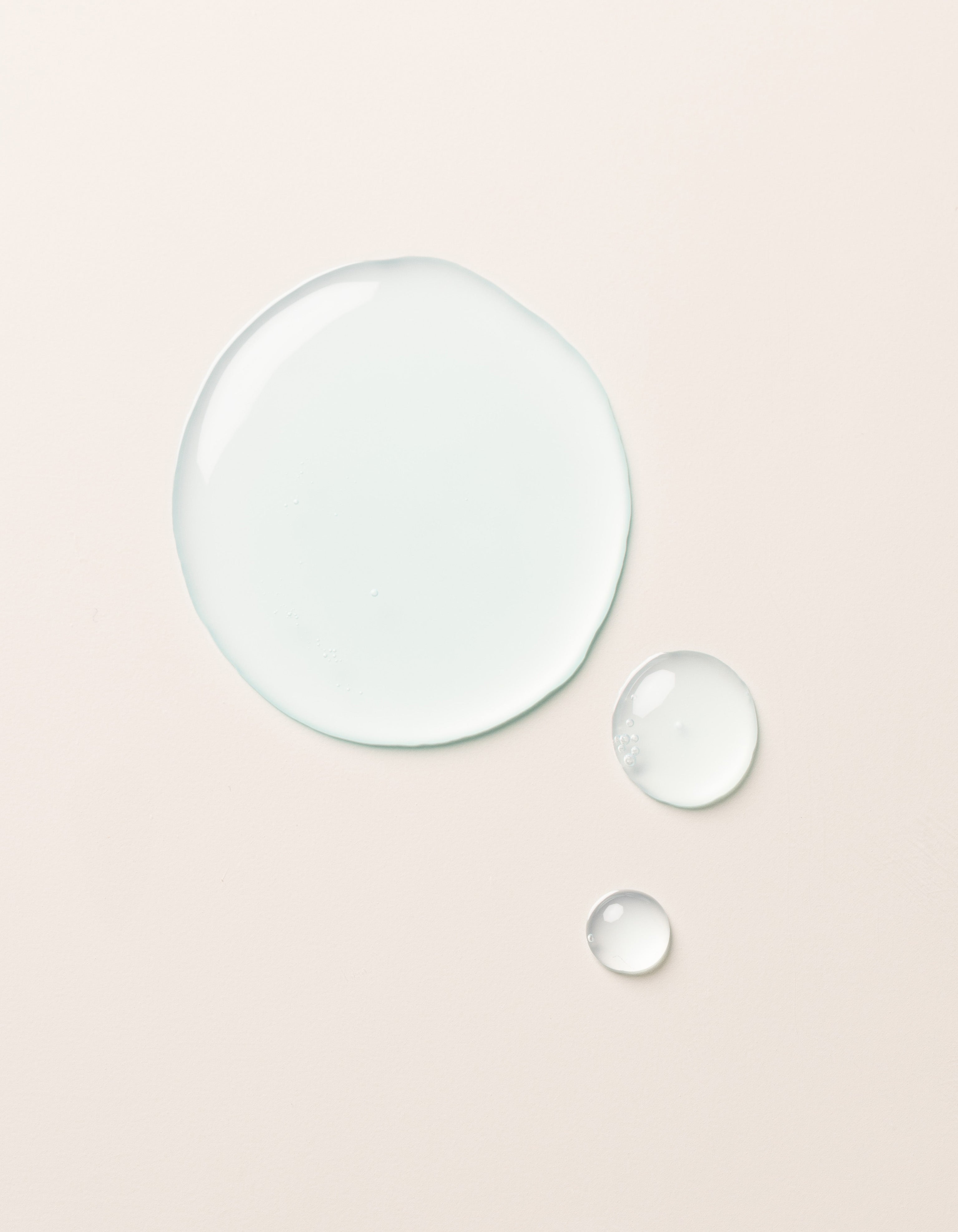 The Hydration Duo by Cozy Earth is shown as a large drop and two smaller drops of clear, light blue liquid on a smooth, light beige surface. The drops vary in size but all maintain a rounded shape, appearing glossy and slightly reflective.